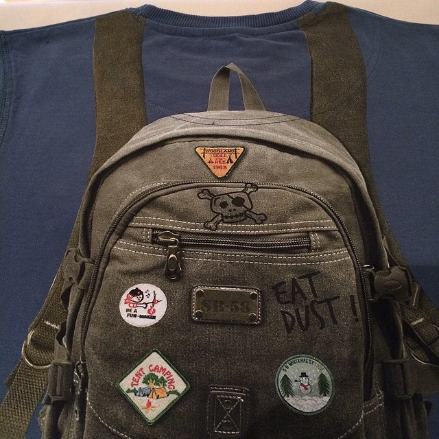 A well-worn olive green backpack adorned with various patches and a skull design rests against a dark blue garment.