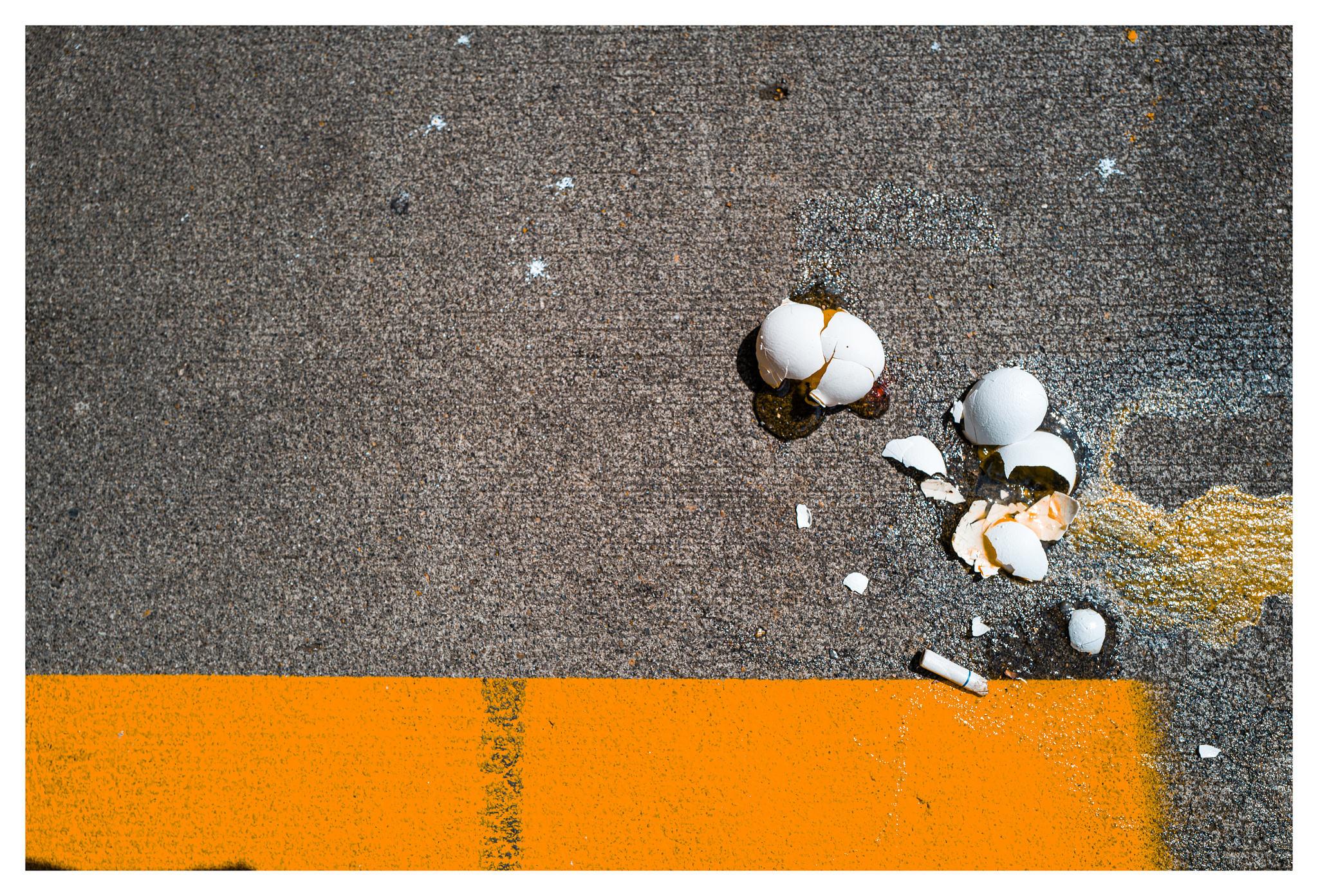 Eggs smashed on a sidewalk over yellow paint and cigarette butts. 