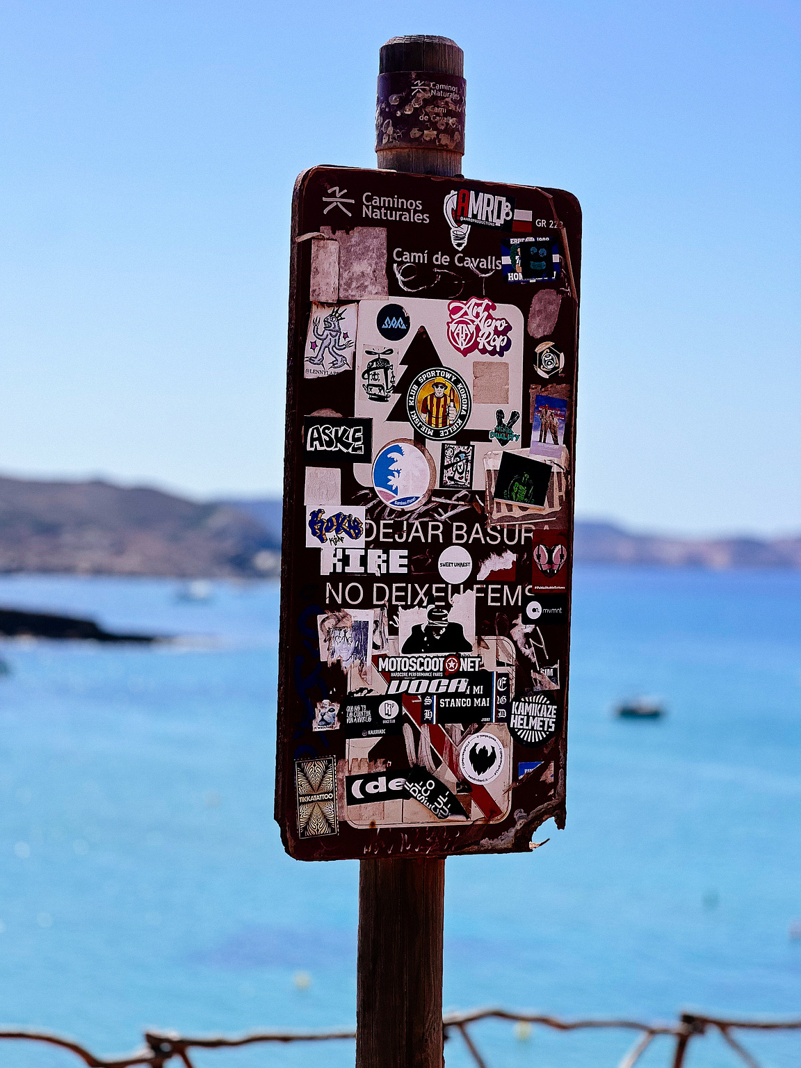 A sign with a lot of stickers, by the sea.