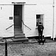 A street scene looking at a whitewashed cottage that has steps leading up to an entrance door on the left, and on the right hand side a window with 'Brocante' written on it. In front of that on the cobbled street, as if waiting for someone inside the Brocante shop, stands a man with dark clothing and a beanie type hat, his hands clasped in front of him, holding a dog on a lead. on a lead.