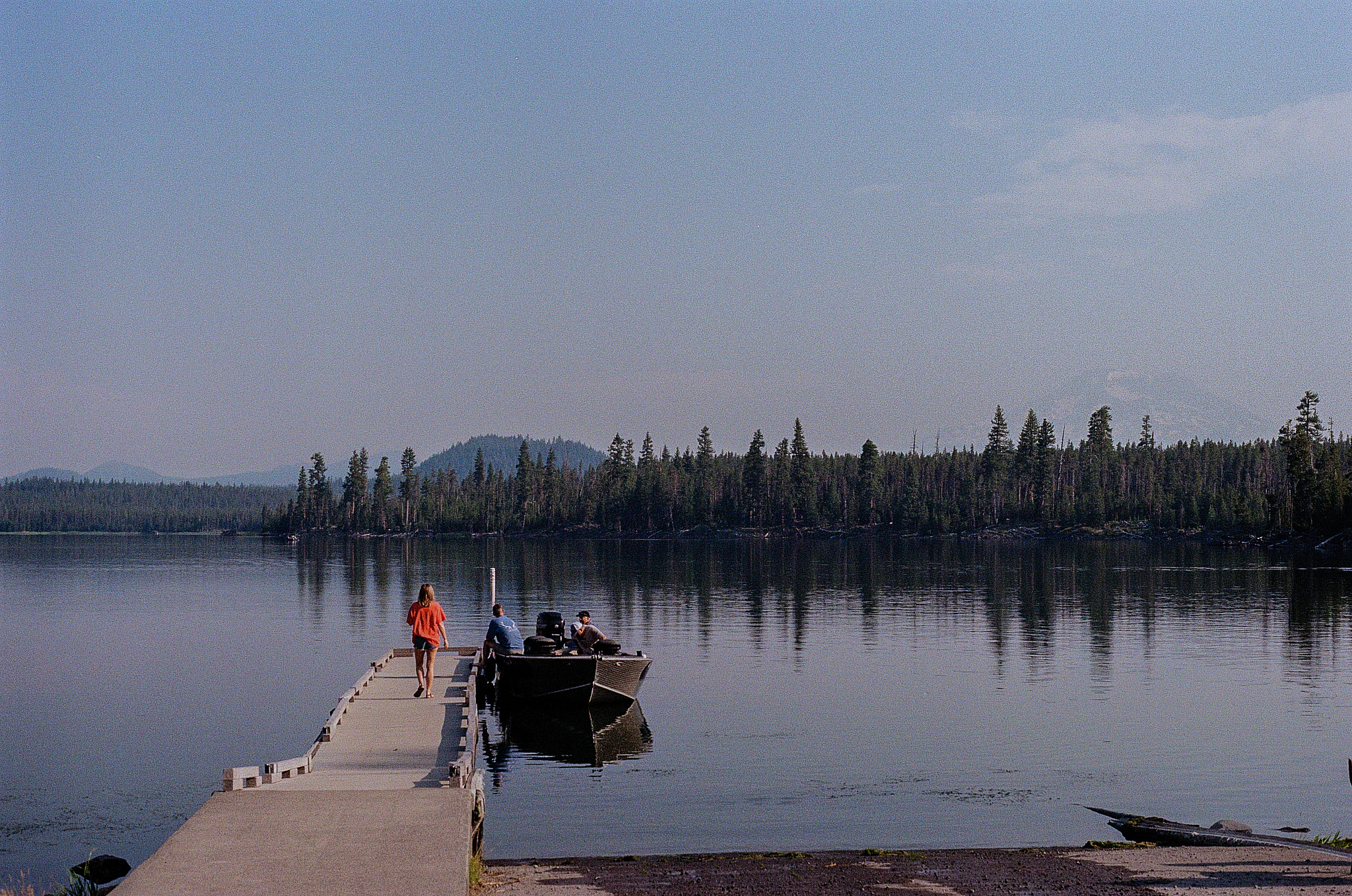 A girl in a bright orange shirt walks toward a boat on a long dock on a mountain lake. 