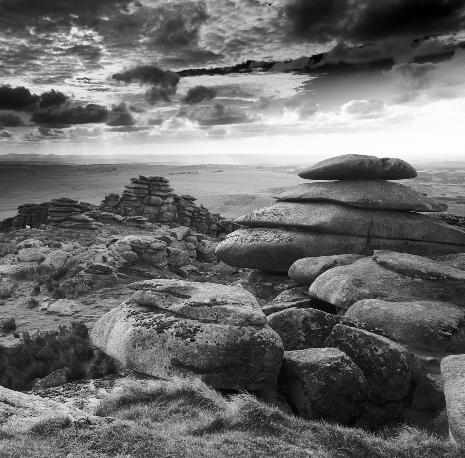 Black and white, piles of granite rocks, weathered and eroded over millenia to look like they're arranged in stacks and piles in the foreground, with a far reaching moorland landscape stretching beyond them, a dark and threatening looking cloudy sky above them, with rays of sunlight poking through in places in the distance...