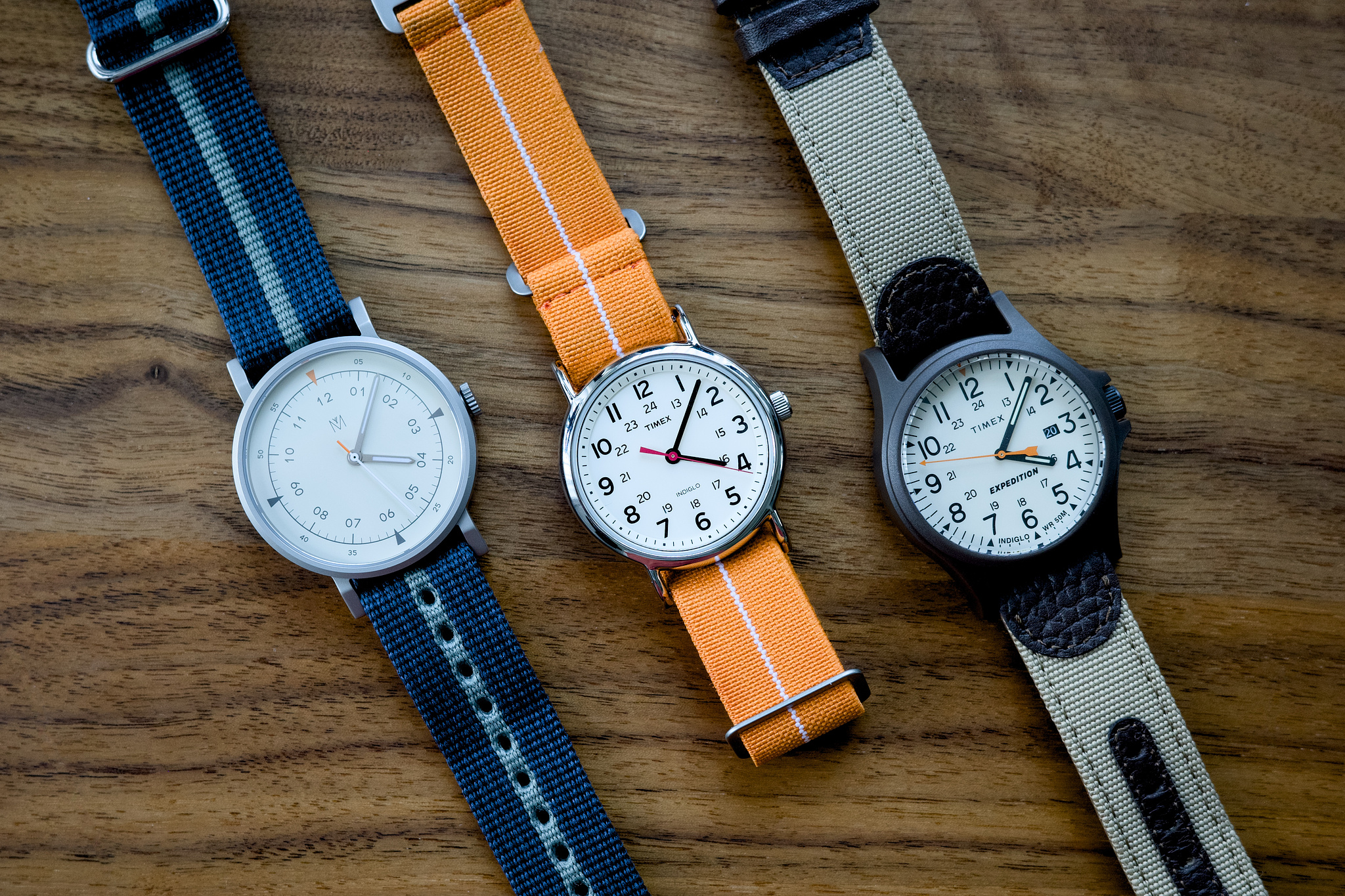 Three mechanical watches, a Maven MUS-01, Timex Weekender, and Timex Expedition Arcadia, are laid out on a walnut desktop. They're arranged side-by-side at a 45 degree angle so their straps look like stripes passing across the image, with the dials centered in the middle of the frame.