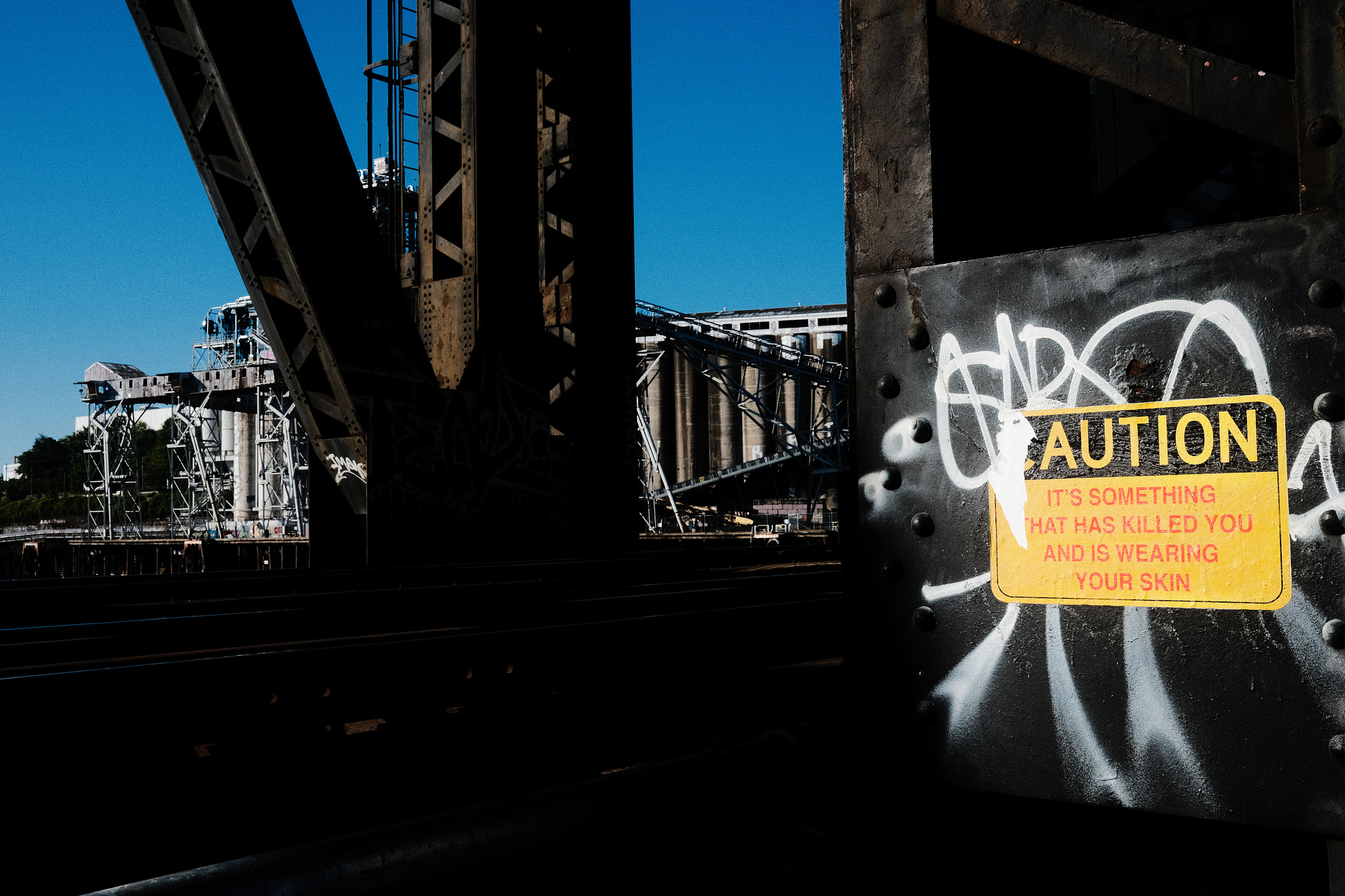 A sign wheatpasted to a steel girder that reads "CAUTION: It's something that has killed you and is wearing your skin."