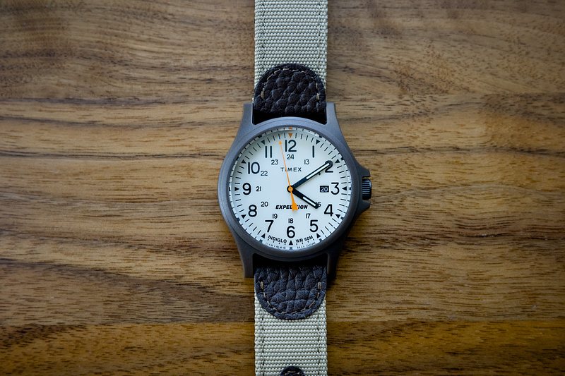 A Timex Expedition Acadia watch sits on a walnut desktop, with a canvas & leather strap.