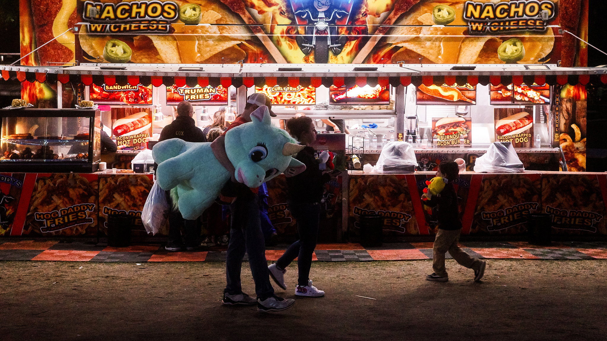 A man carrhing a big blue stuffed unicorn in front of a hotdog stand on a carnival midway. 