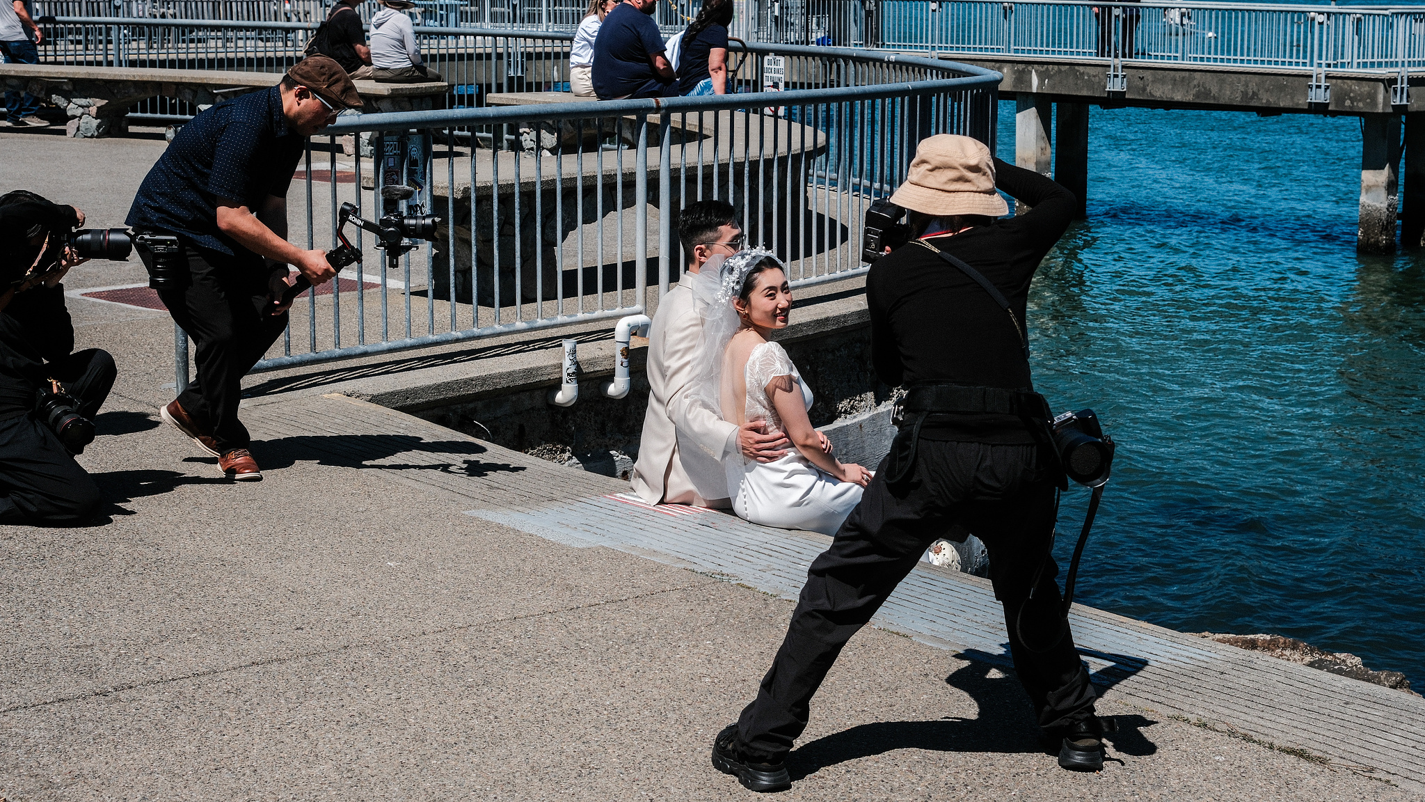 Three people photographing a wedding couple on the sidewalk by a pier