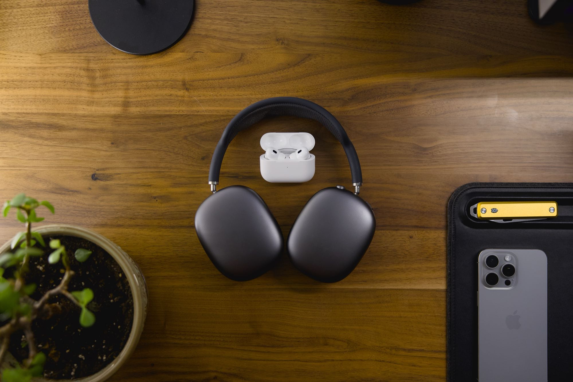 Flat-lay featuring AirPods Max and AirPods Pro 2 sitting on a walnut desktop, with a plant, iPhone, and pocket knife arranged around them.