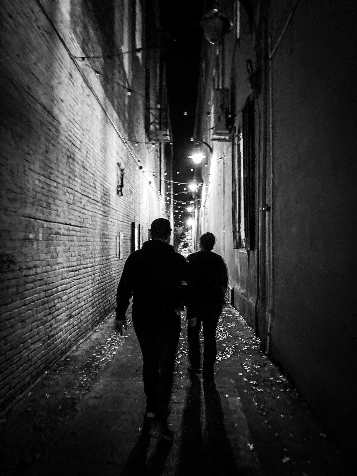 Silhouette of two people walking down a dark alleyway towards bright street lights.