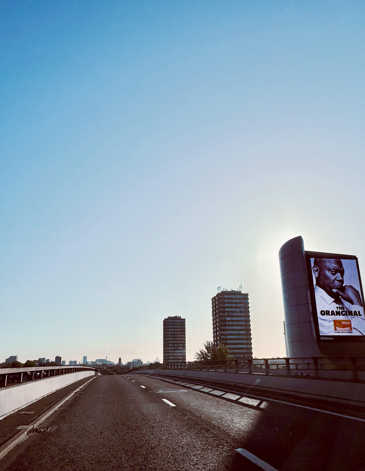 Empty highway with a billboard and twin high-rise buildings against a clear sky, with the sun just above the horizon.