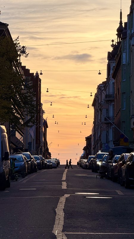 Das Bild zeigt eine Straße bei Sonnenuntergang mit Autos, die auf beiden Seiten geparkt sind. Die Silhouette von zwei Personen ist in der Ferne sichtbar. Der Himmel ist in warmen Gelb- und Orangetönen gefärbt, was eine ruhige Atmosphäre schafft.