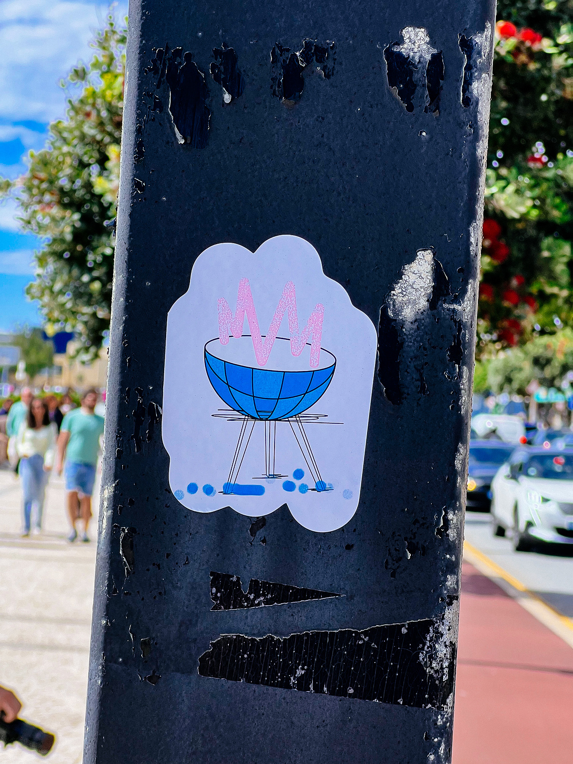 A sticker depicts a blue bowl with pink foam on a light pole, textured with peeling layers, against an unfocused urban backdrop.