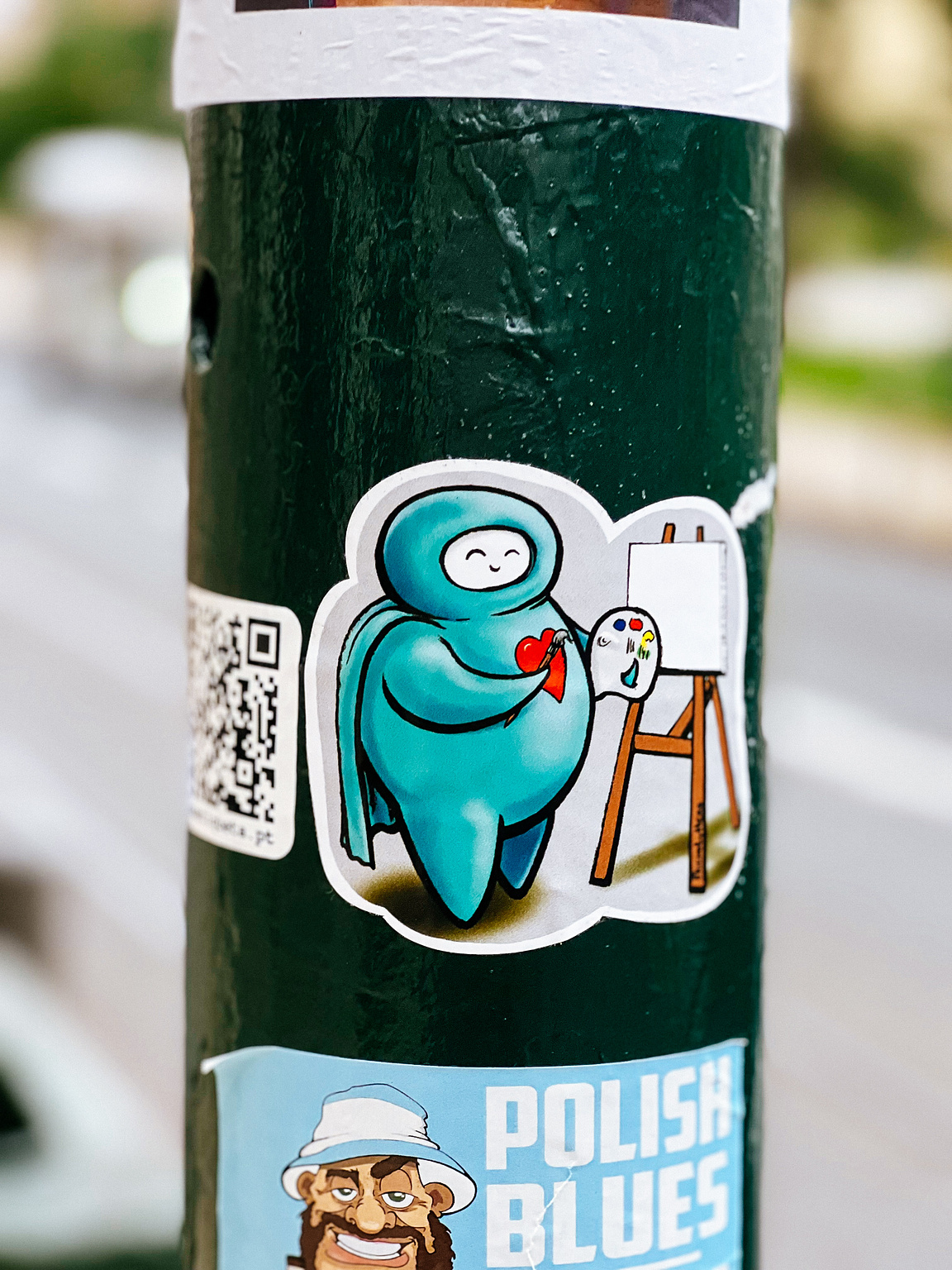 Sticker of a blue character painting on an easel, affixed to a textured green pole with other stickers and a QR code visible.