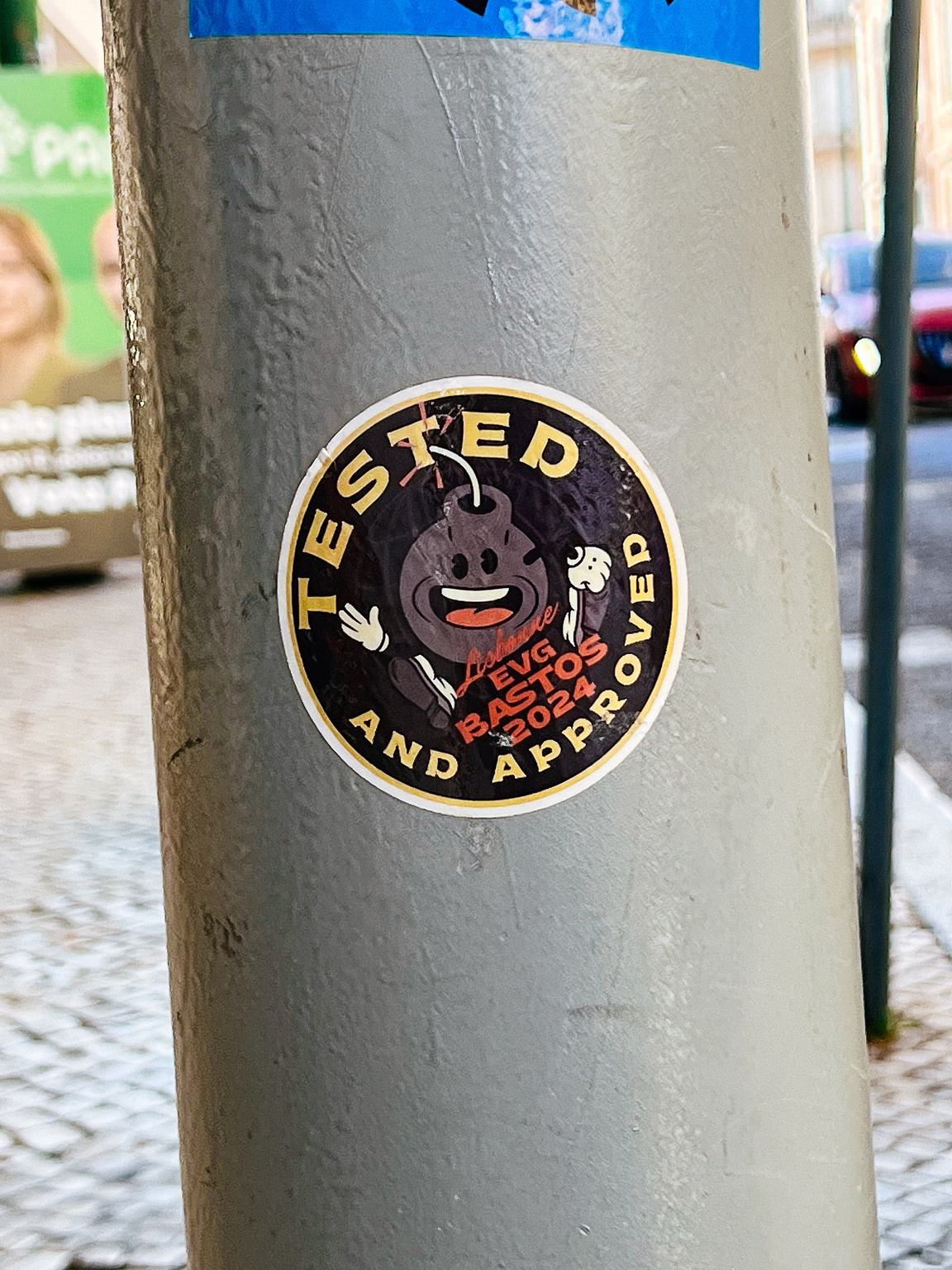 A sticker with a cartoonish bomb character thumbs up, says "TESTED AND APPROVED" on a grey pole, urban background.