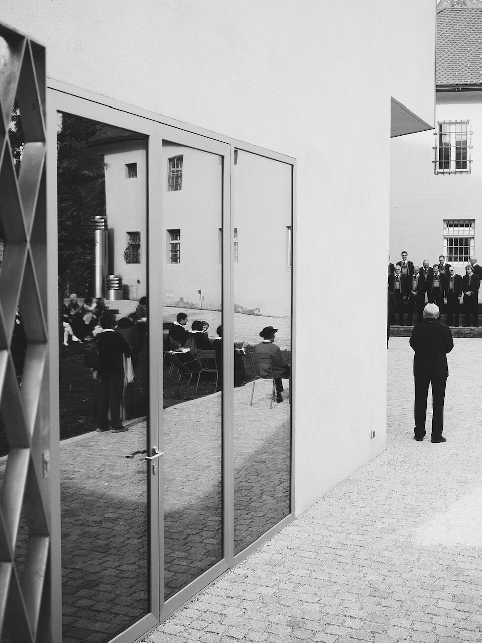 A black and white photo showing an outdoor gathering. In the foreground, a large glass window reflects people sitting on chairs, with one person standing nearby. In the background, a group of people stands near a building with barred windows. The scene suggests a formal or ceremonial event, possibly a celebration or presentation.