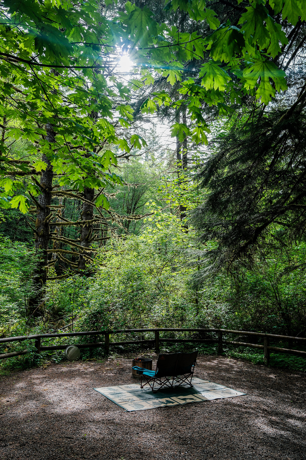 A folding love seat on a green mat in front of a silver Solo Stove under trees. 