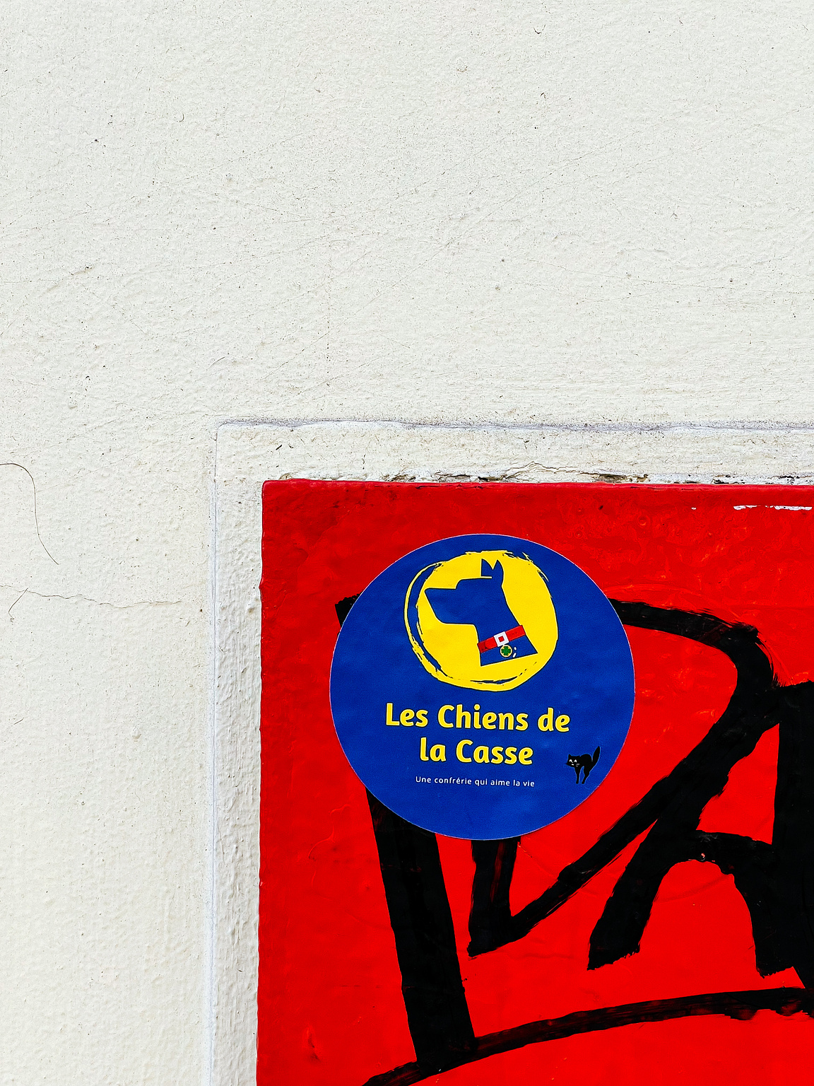 A sticker on a wall with red graffiti depicts a dog inside a yellow circle, text reads "Les Chiens de la Casse, Une confrérie qui aime la vie".