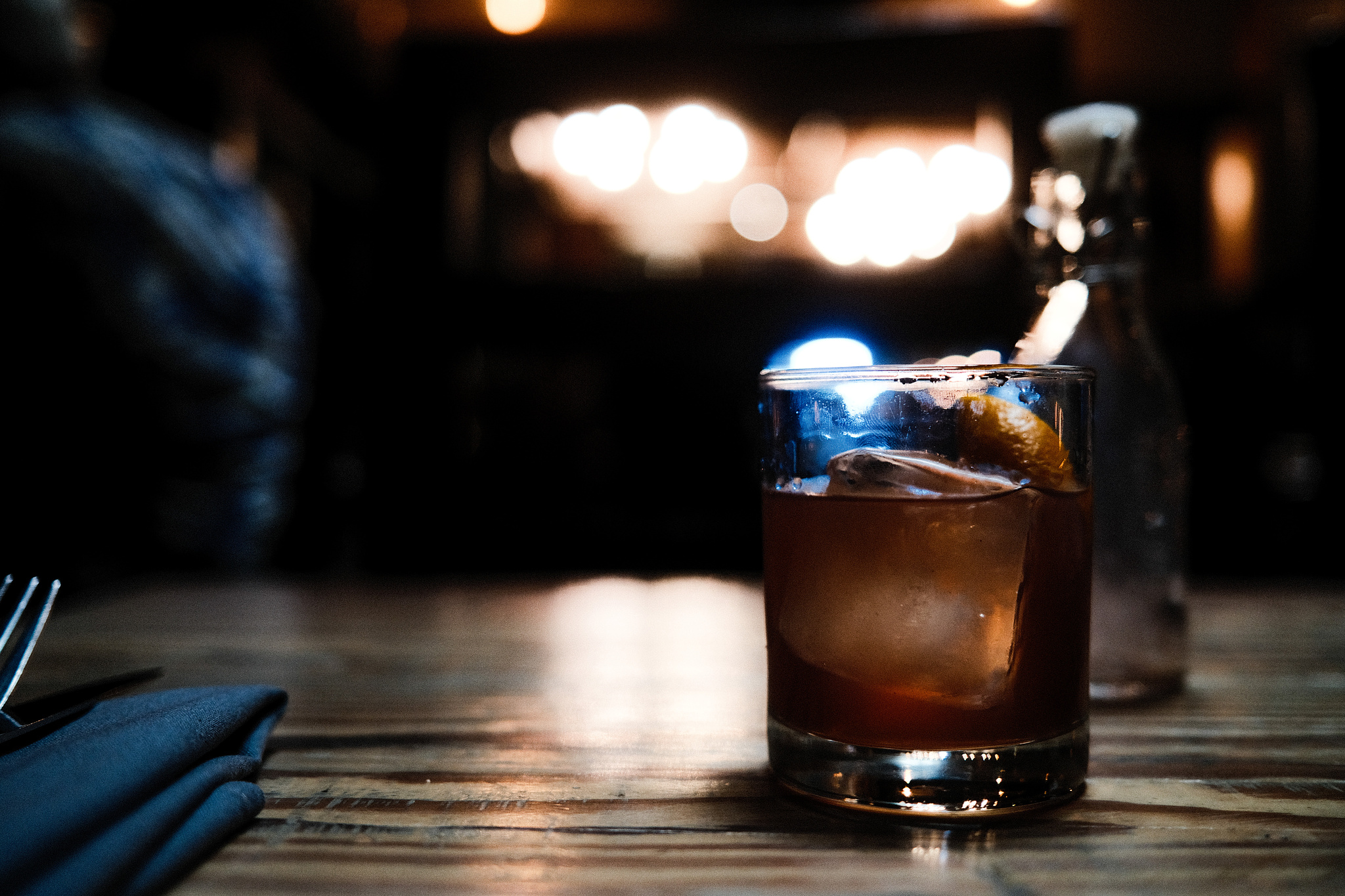 A glass of whiskey with a large square cube and a twist. The little jar it was poured out of in the background. 