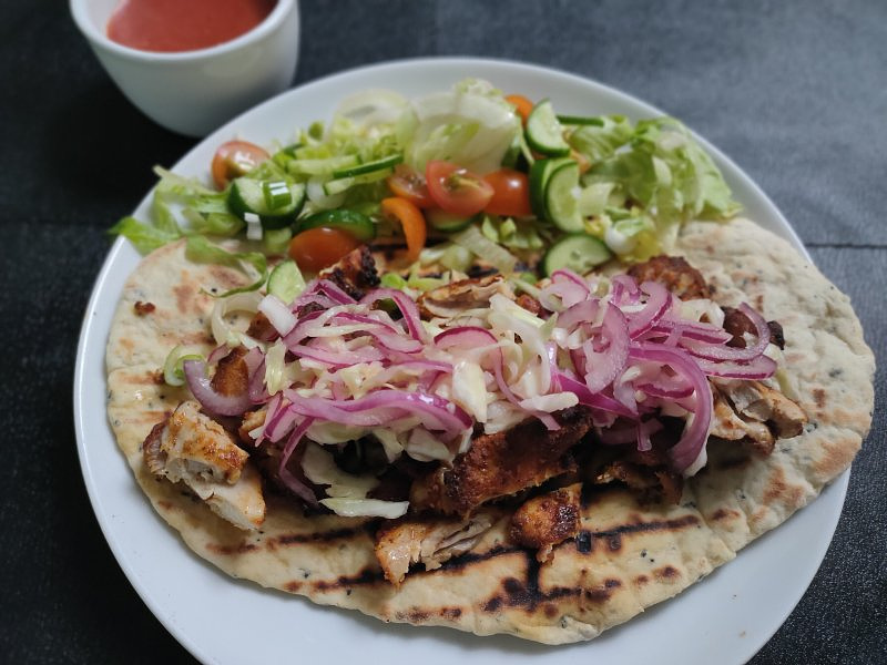 Photo of white flatbread, with bbq cooked chicen, red onion, white onion, salad.  On a white plate.  A small bowl of sauce at the top of the image.