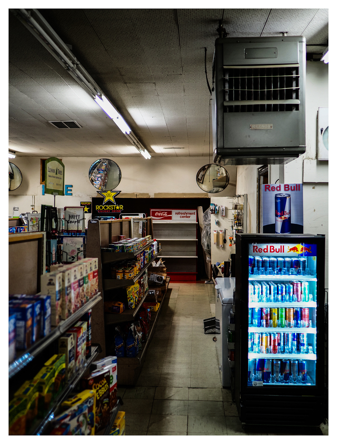 A Red Bull cooler case glows under a half-lit country store aisle