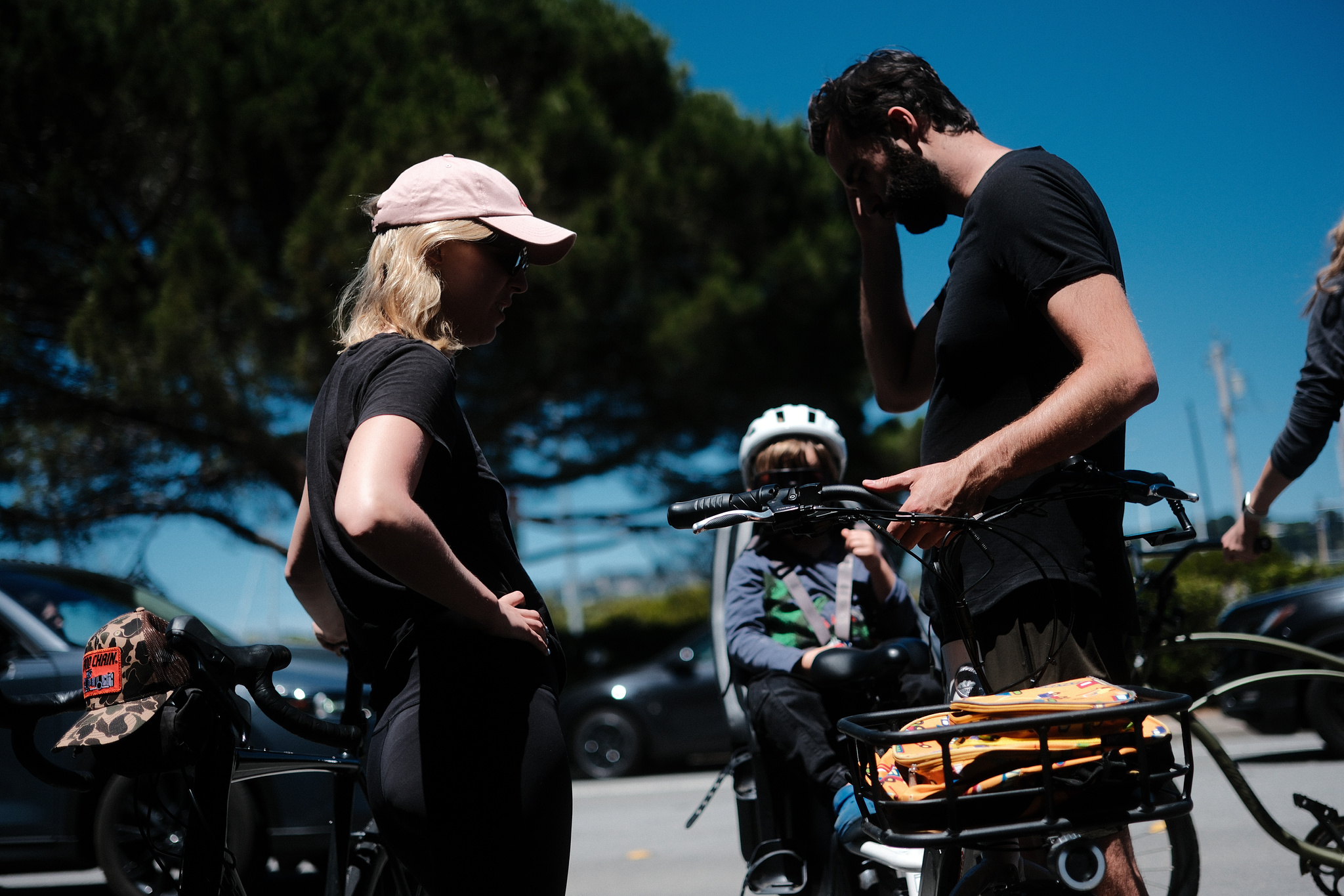 Young parents struggle with a young child taking his time to settle into his bicycle seat. 