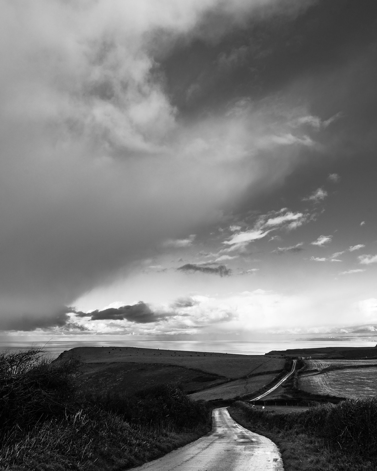 Portrait format black and white landscape. A coastal scene with a wet single track lanes winds throught the rolling clifftop countryside into the distance at the bottom of the frame, with a calm glassy sea just over the cliffs. The majority of the frame is taken up with a sky puntuated with cumulus cloud on the right but with a big stormy looking one coming into the frame from the left.