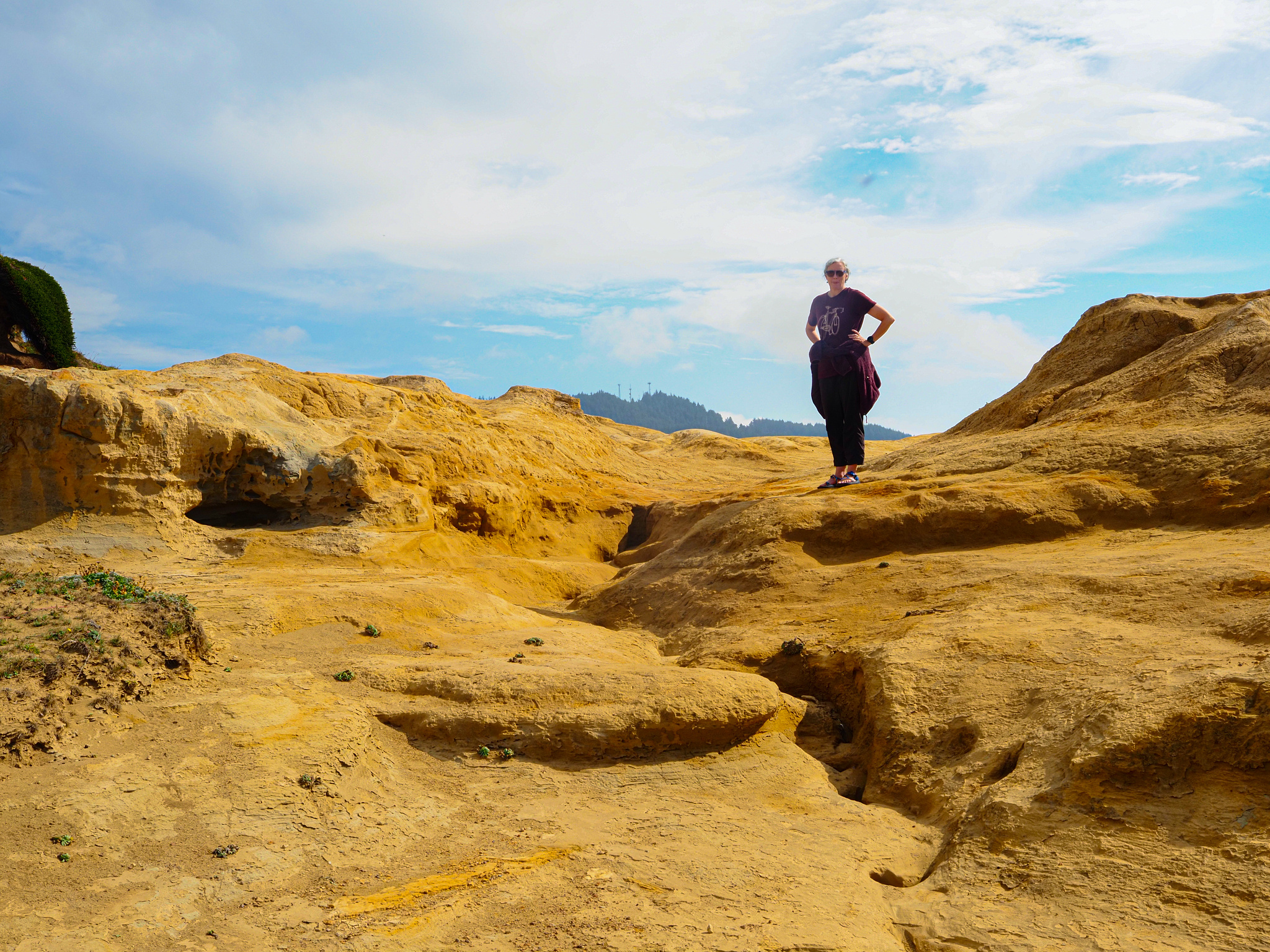 A woman stands on orange rock and dirt with blue and cloudy  skies behind her