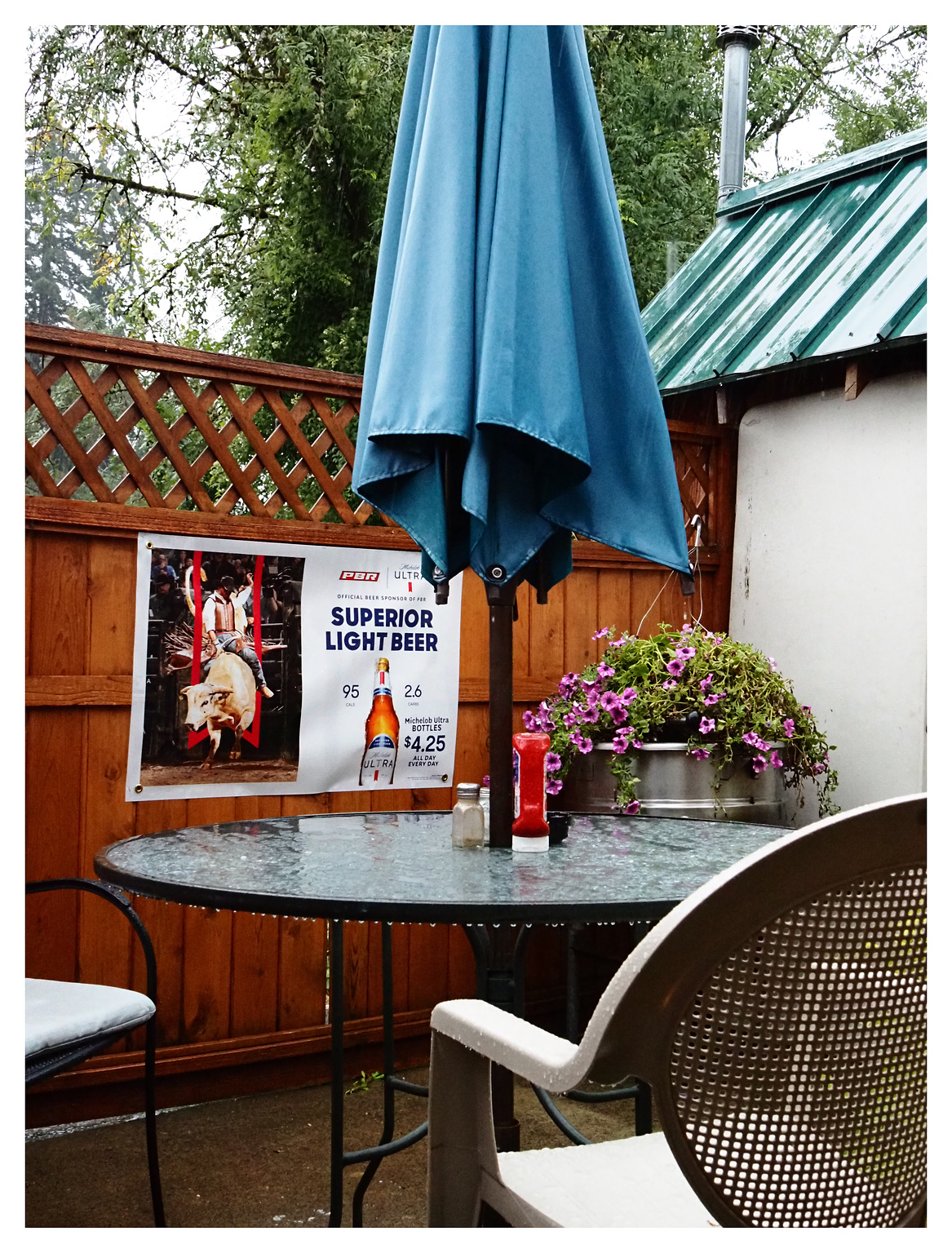 An umbrella table with a blue umbrella, wet in the rain. Beer sign on the wooden fence behind it 