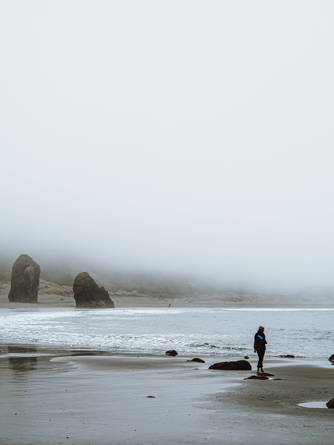 A woman walks along a foggy beach, two tumbled rocks obscured by mist in the background