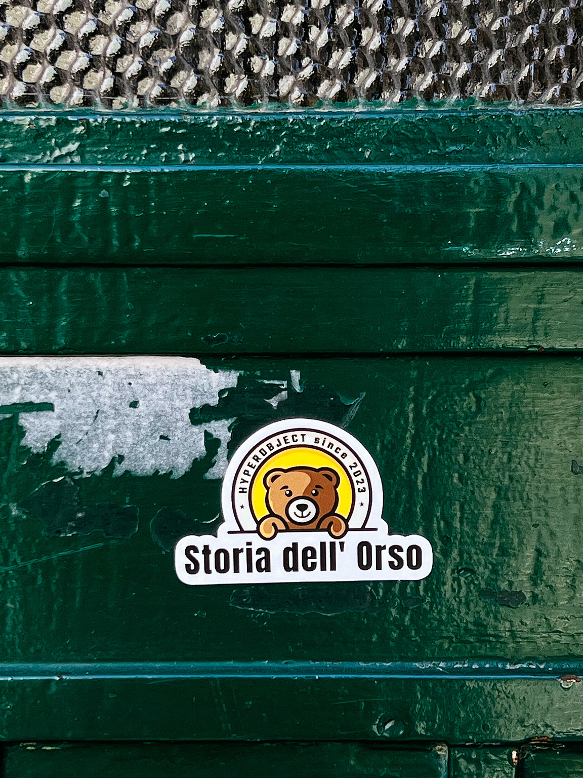 A sticker with a cartoon bear is adhered to a green, textured surface.