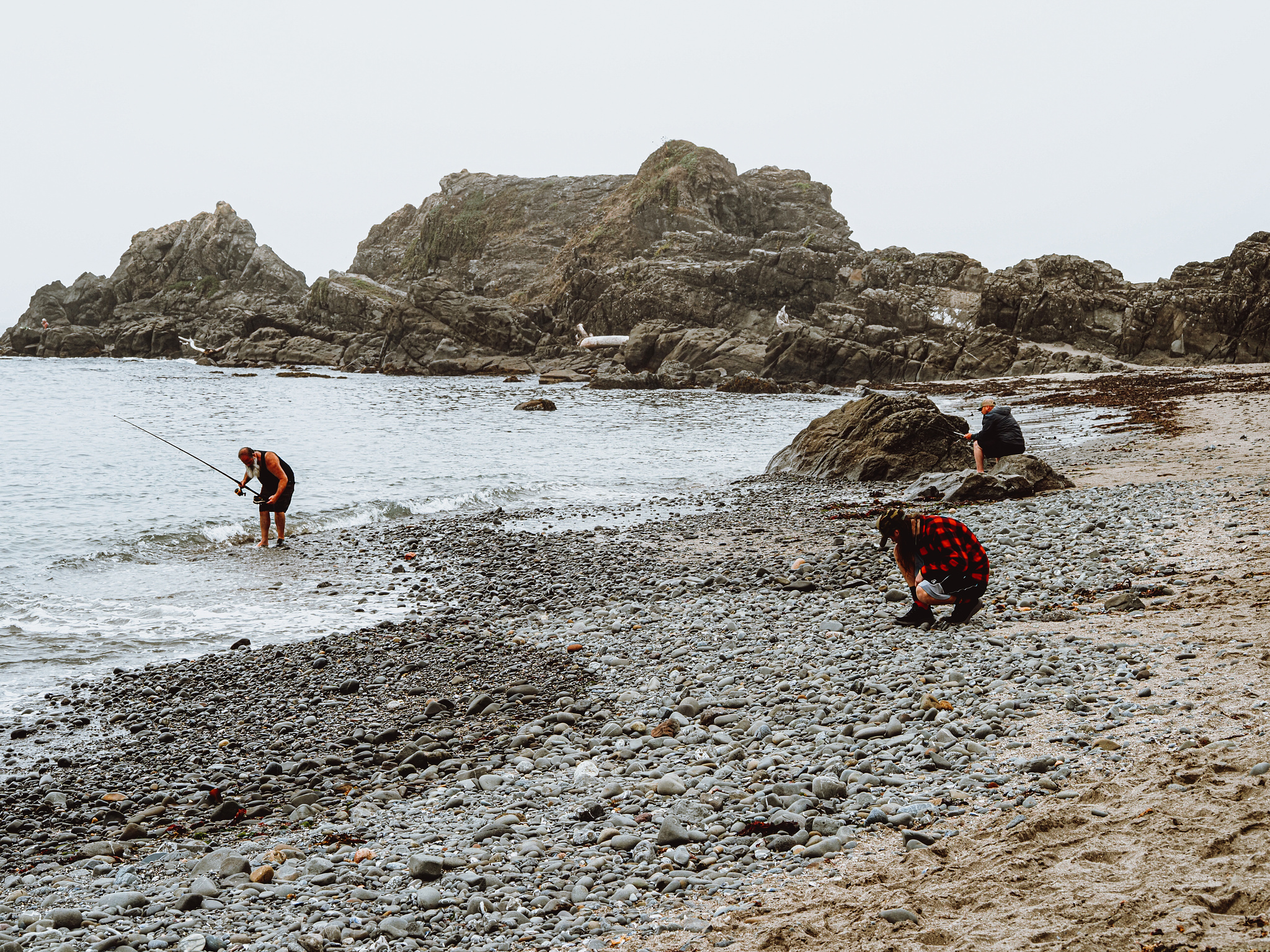 A fisherman in the water bends down looking at his feet. A man in a red and black check flannel kneels on a rocky beach