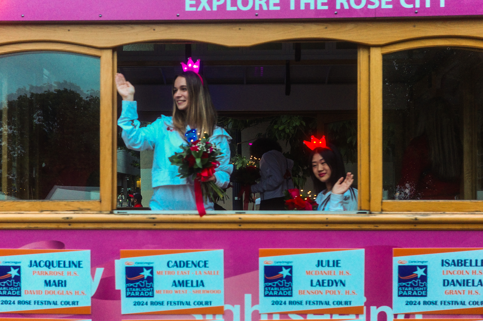 Two young women in tiaras waving from a pink streetcar