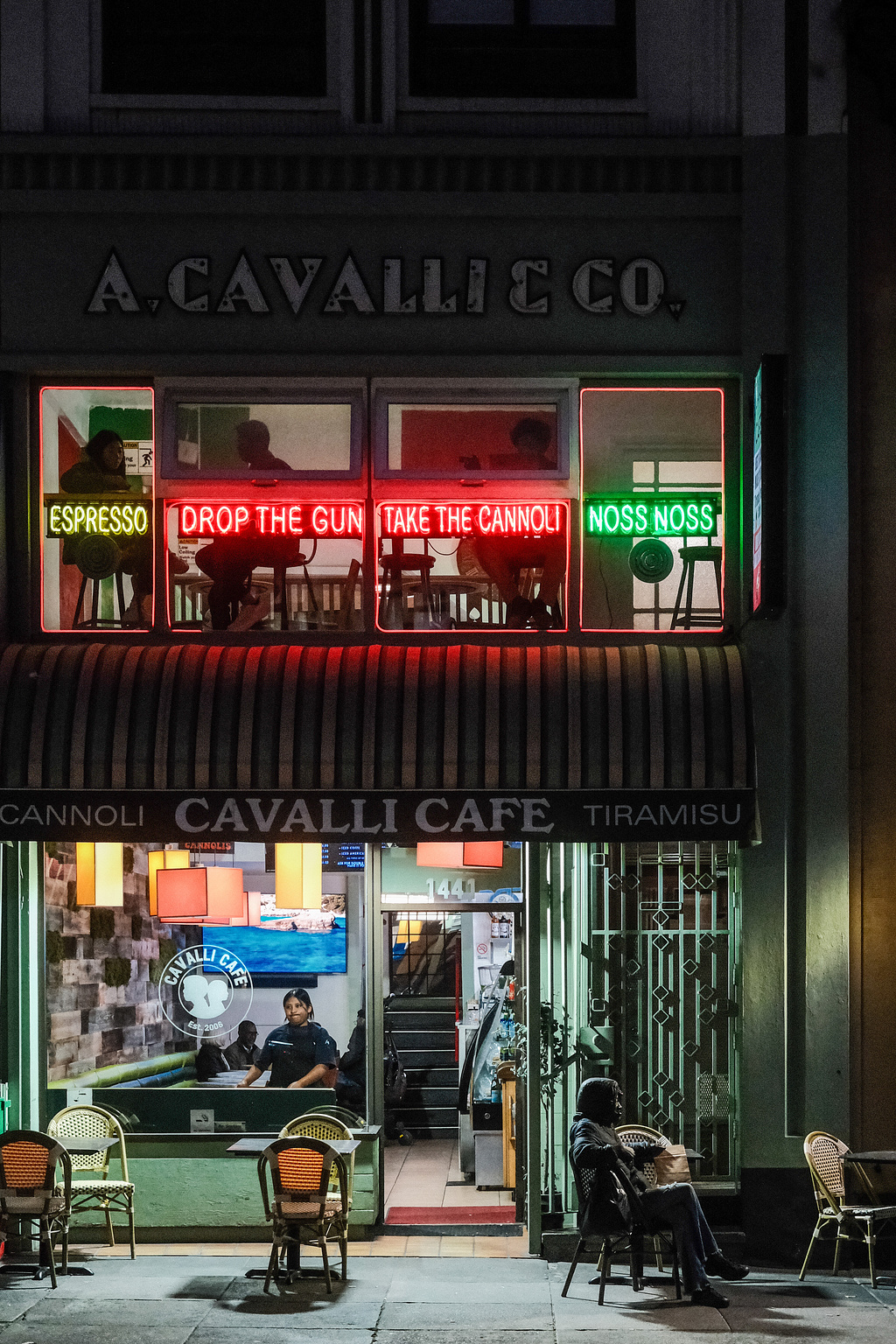 A person sits alone on a dark sidewalk in front of an Italian cafe