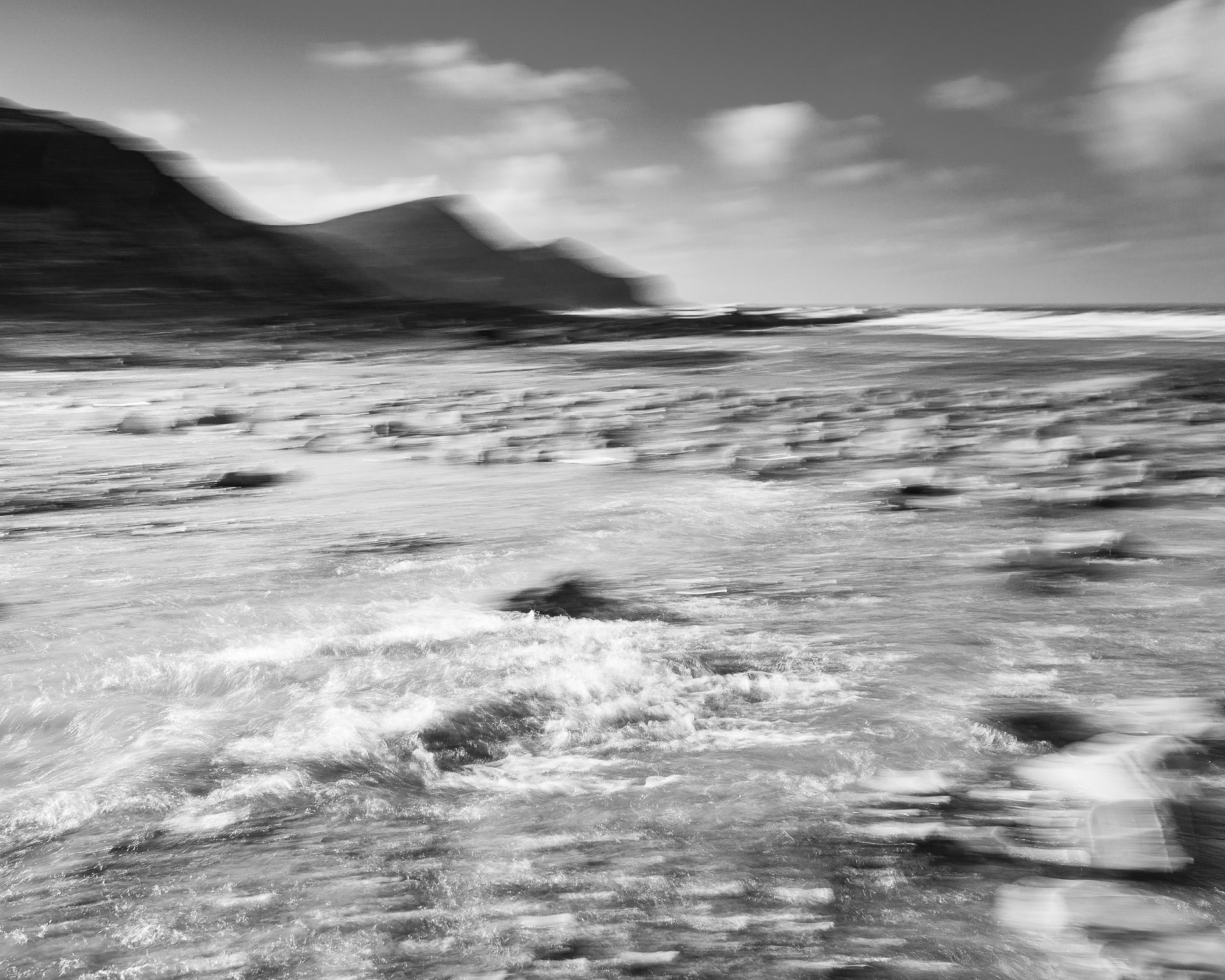 Black and white landscape taken on the turn of the tide from a low angle at a flat beach. The picture is taken with a slow shutter speed handheld, resulting in blurred movement. There is a shallow white wave in the foreground, with water running over submerged rocks behind, and receding out to a headland and the horizon with a mostly clear sky and a few fluffy clouds above.