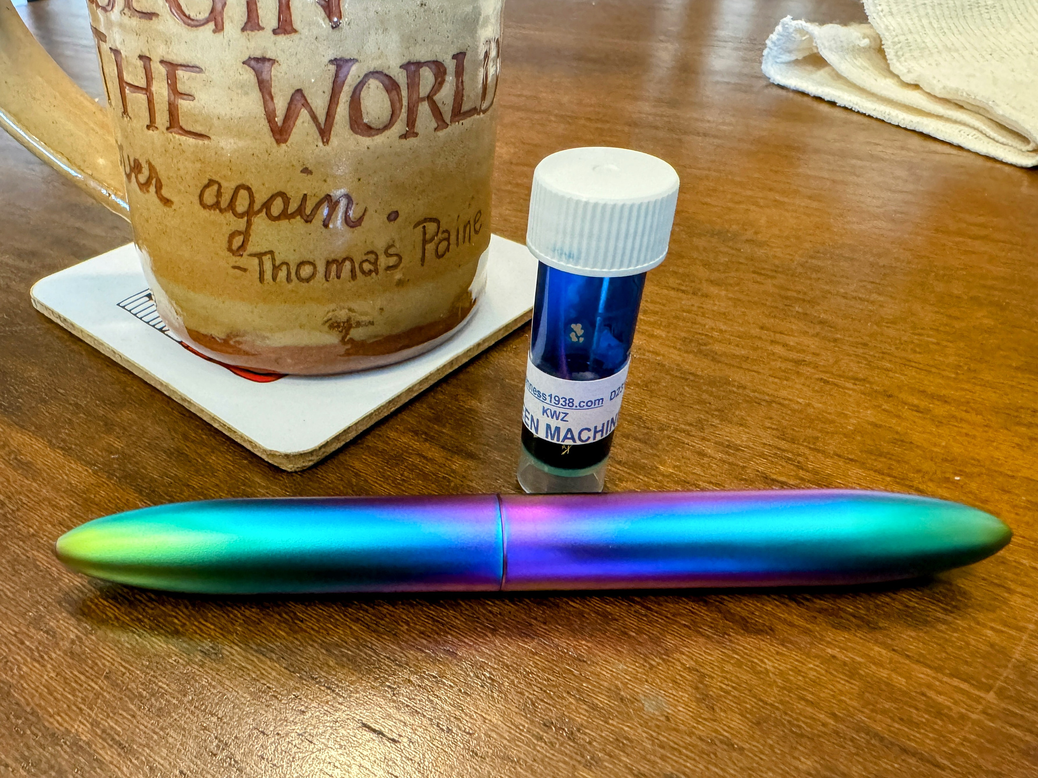 Multicolored metal fountain pen with small ink sample vial and part of a coffee mug visible