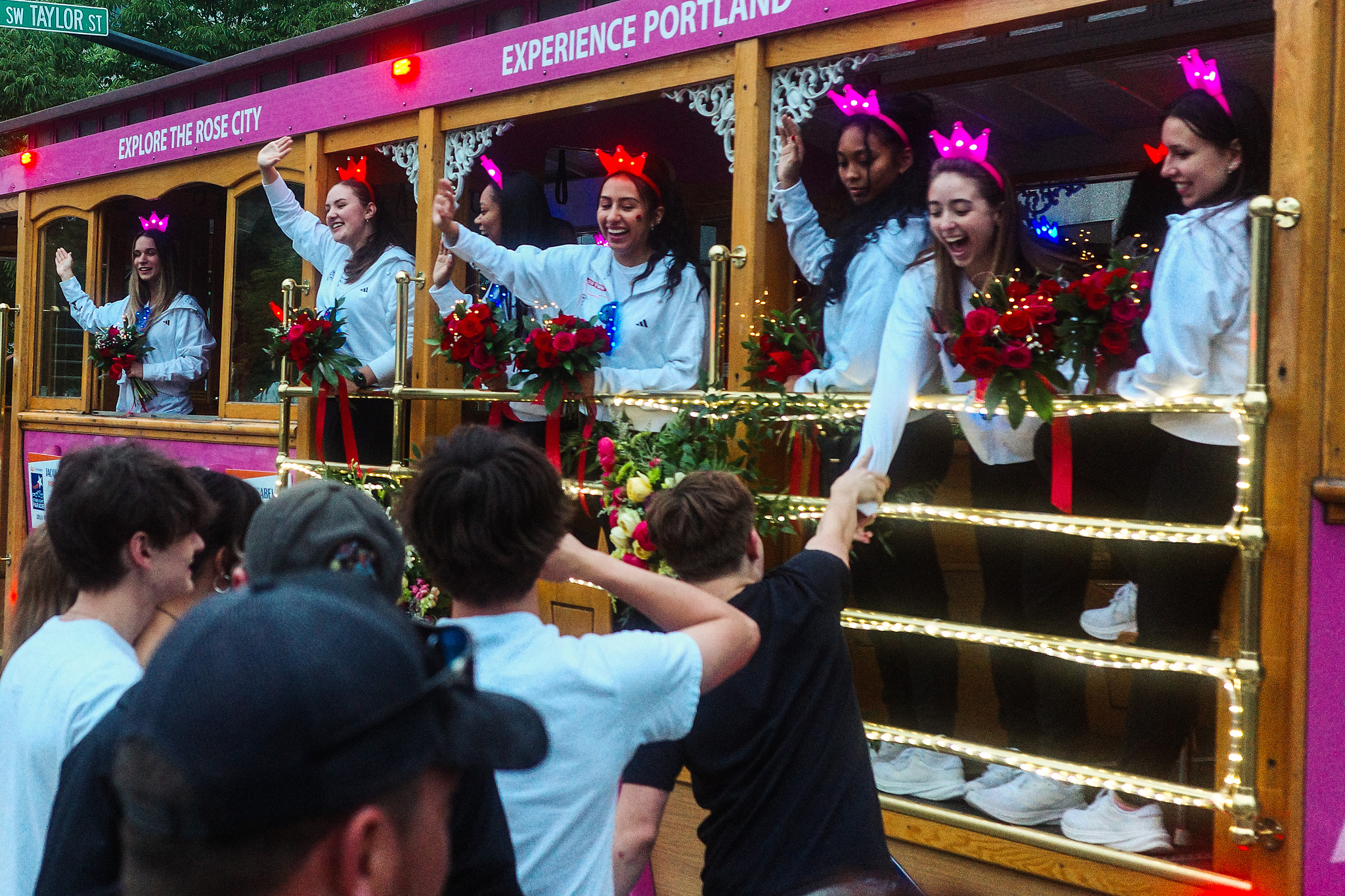 Young women in a street car smile and wave. One of them is reaching to take the hand of someone in the crowd on the street. 