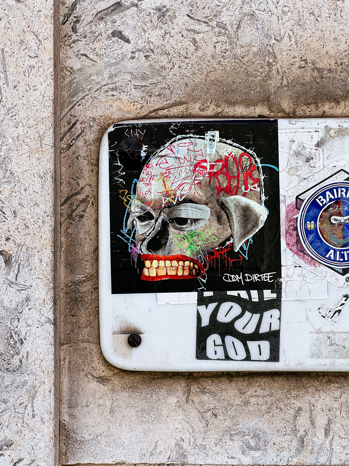 A decal of a skull with graffiti on a utility box against a textured wall, with the phrase "KILL YOUR GOD" below it.