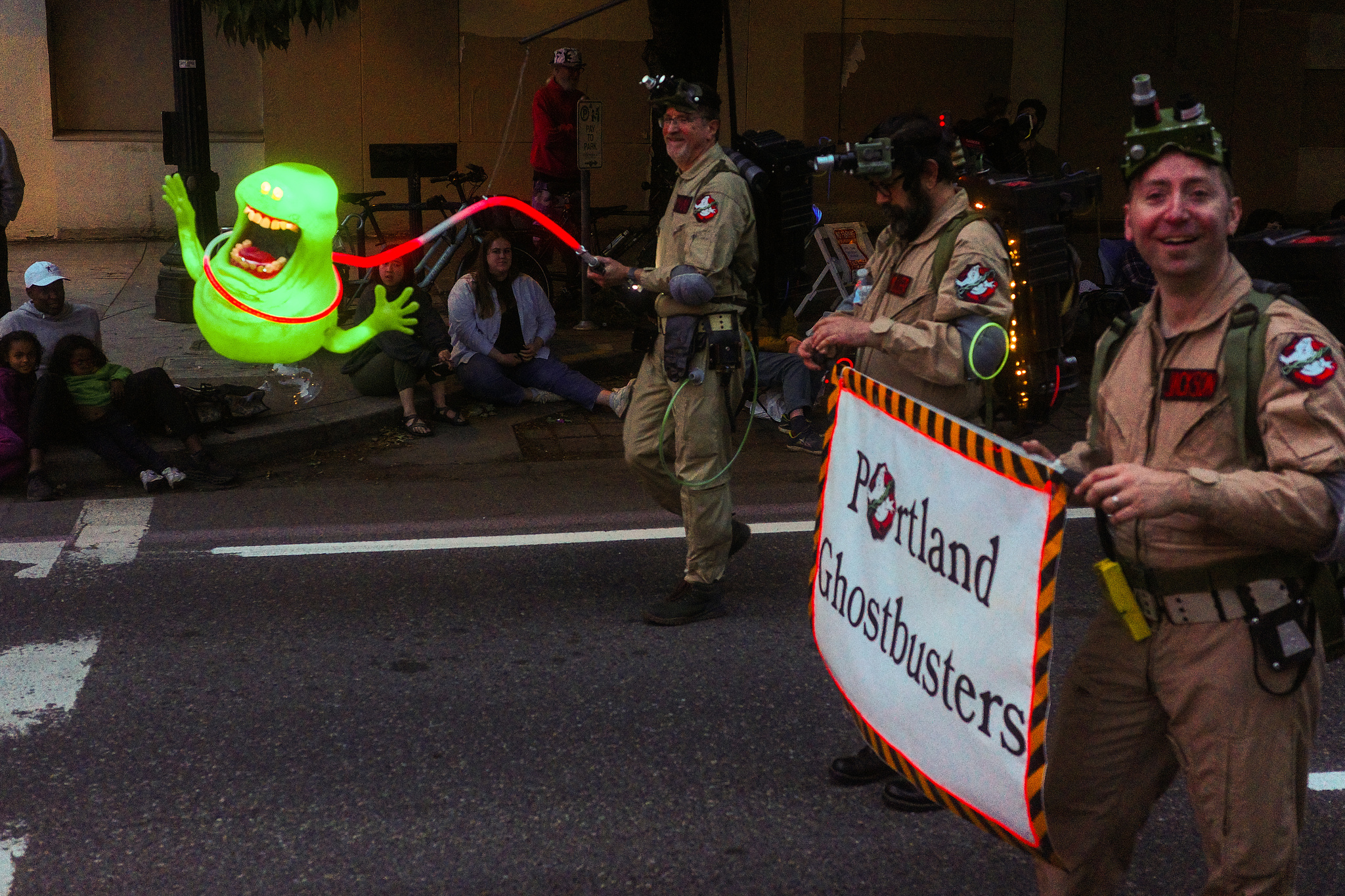 People in Ghostbuster uniforms marching in a parade. One of them has a Slimer on a glowing red leash. 