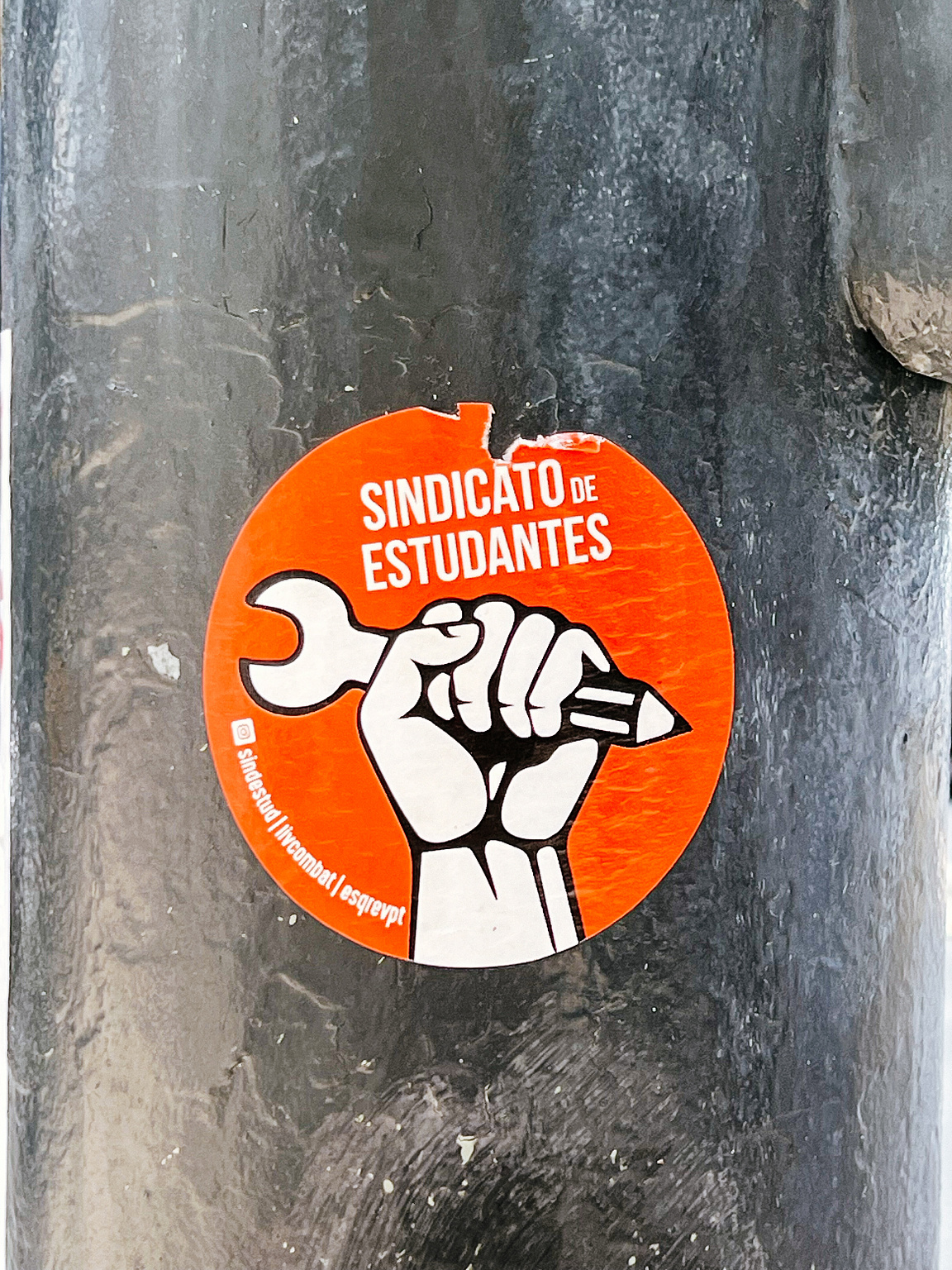 A sticker with a wrench/pencil-clenched fist against an orange backdrop, labeled "SINDICATO DE ESTUDANTES," adhered to a textured, vertical pole.