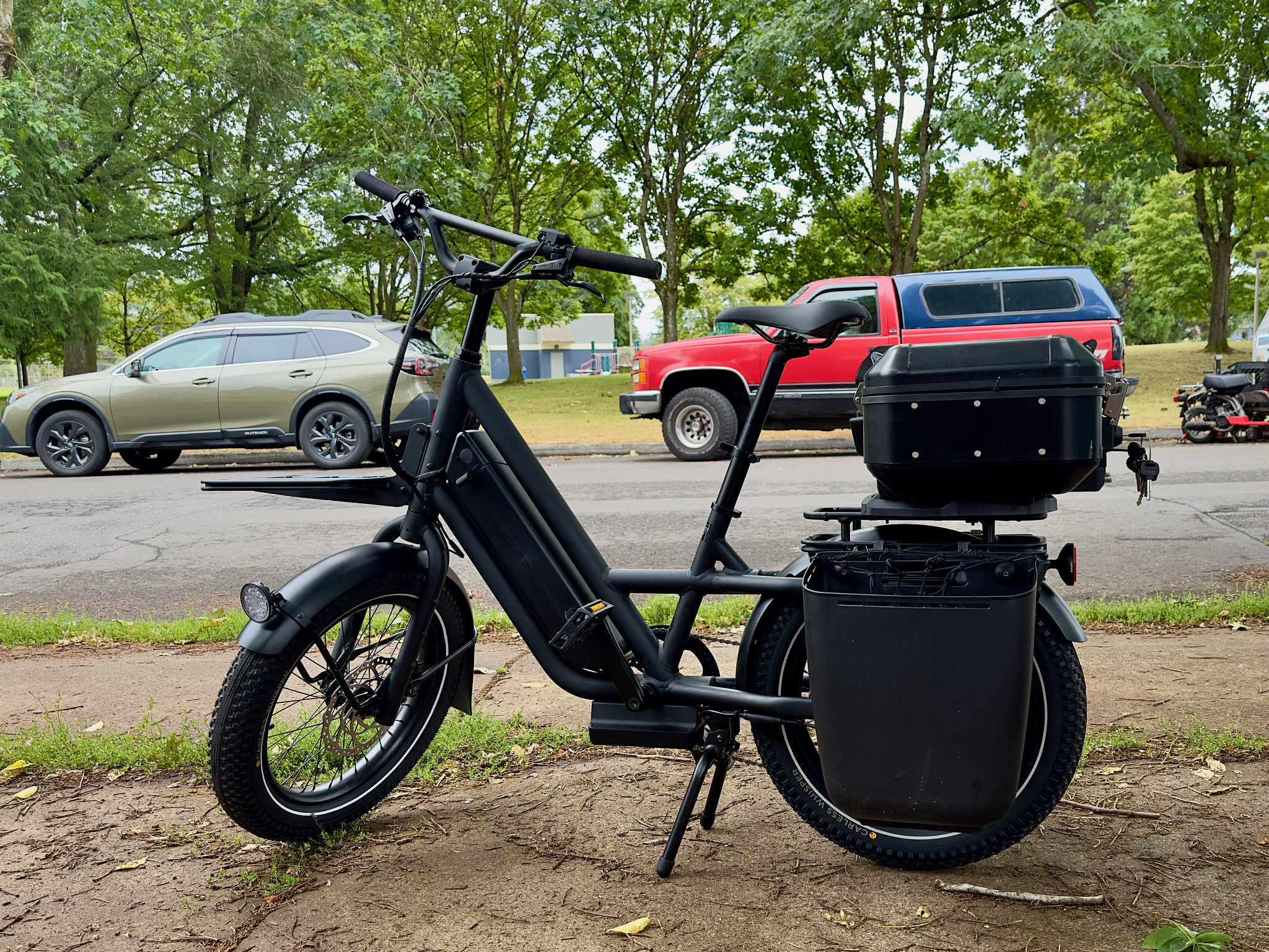 A black short-tail cargo bike with hard panniers on the rear and a motorcycle top case on the rear rack. 