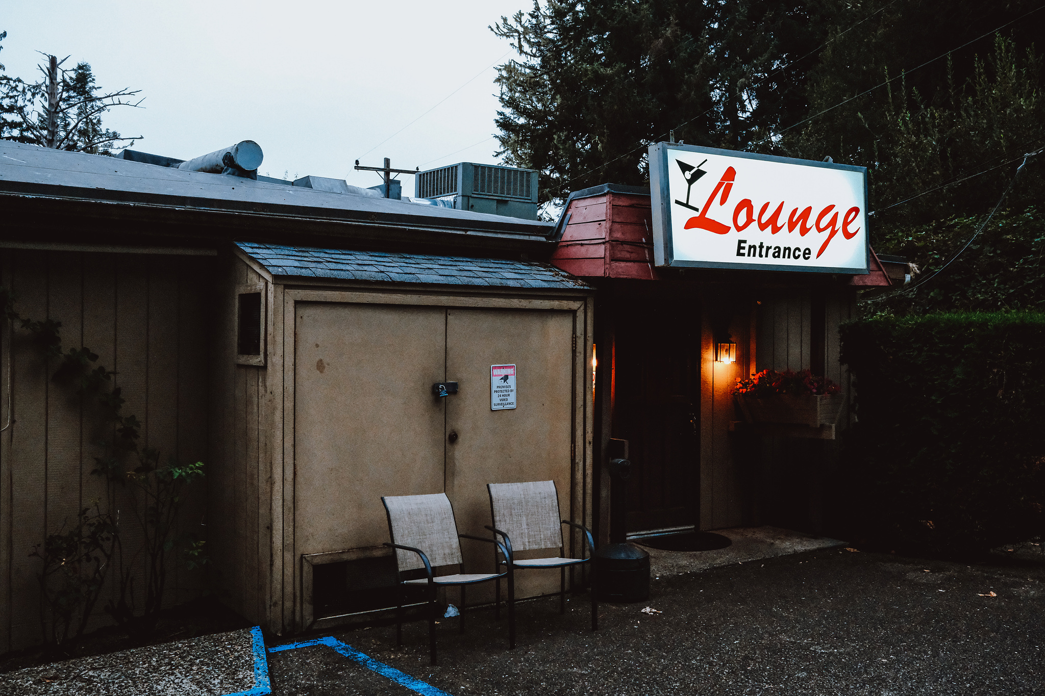 A pair of chairs in front of a run down building under.a white sign that reads "Lounge Entrance" in red lettering with an askew martini glass logo