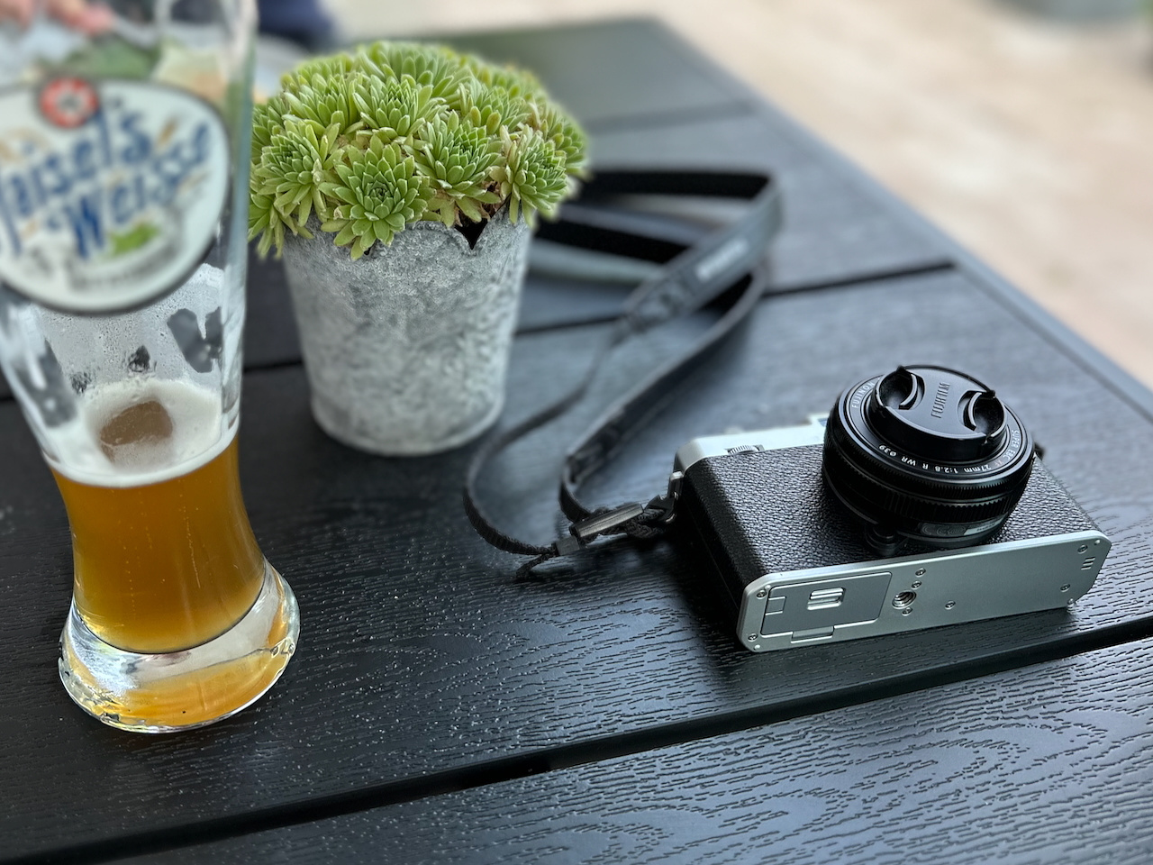 A half-full glass of beer, a potted plant, and a camera rest on a dark wooden table.