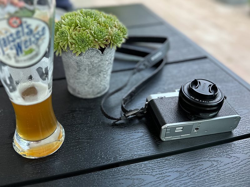 A half-full glass of beer, a potted plant, and a camera rest on a dark wooden table.