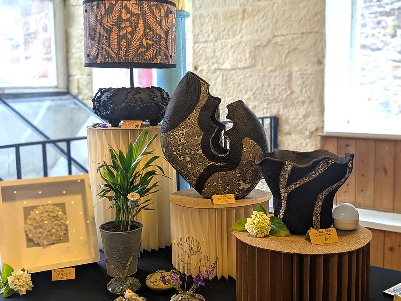Handmade ceramic items, including vases and lamps, on stands in front of the stone walls and windows of an old mill