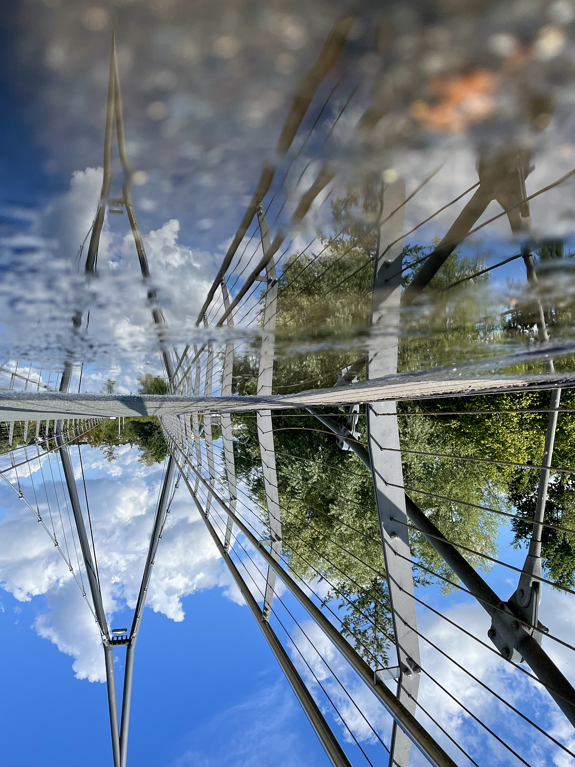Bridge for pedestrians, blus sky with some whhite clouds, reflected in a puddle on the bridge. The whole picture ist turned upsidedown.