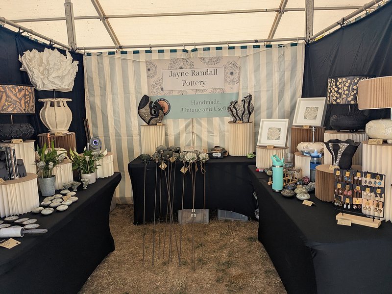 Looking into a shaded exhibition stall, with many ceramic items displayed on tables on the left, right and back in a u shape. There is a water bottle visible on the right stand - it was hot!