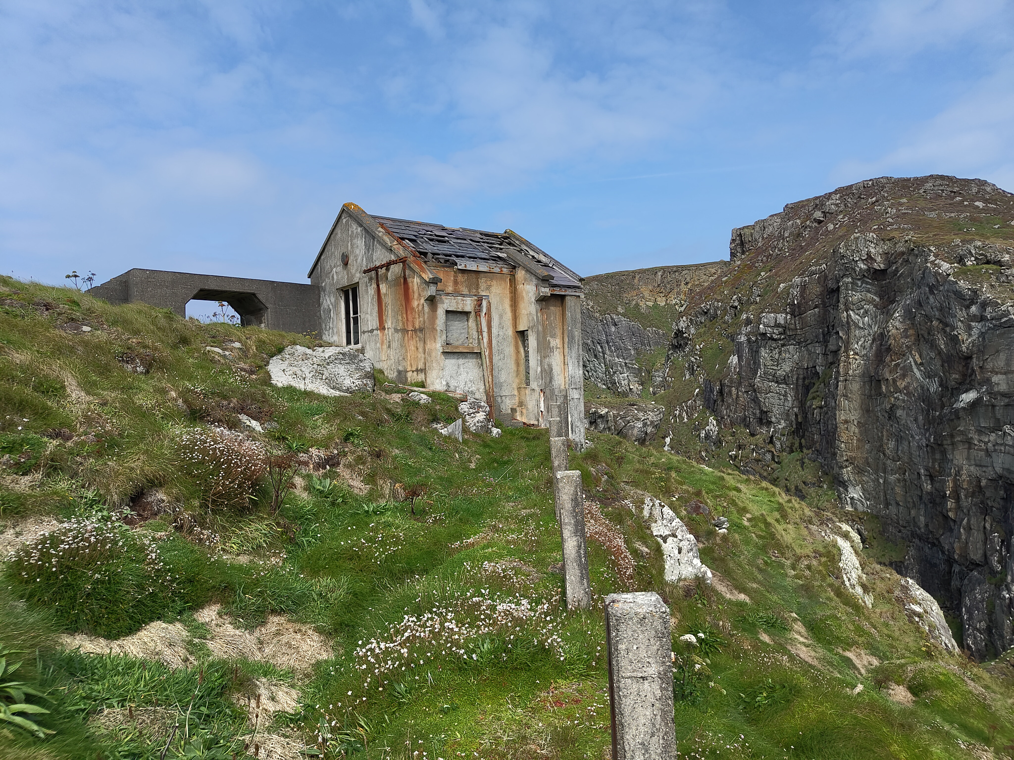 A dilapidated outhouse without most of its roof, covered in rust stains, surrounded by a grassy hill, blue skies and great crags in the background