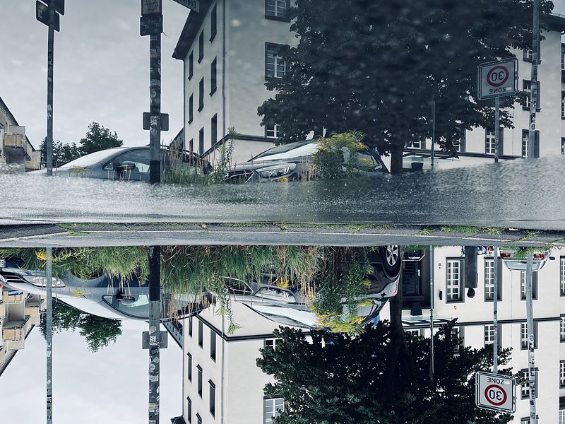 White building, two cars, and a tree, reflected in a puddle on the street. The picture is turned upsidedown.
