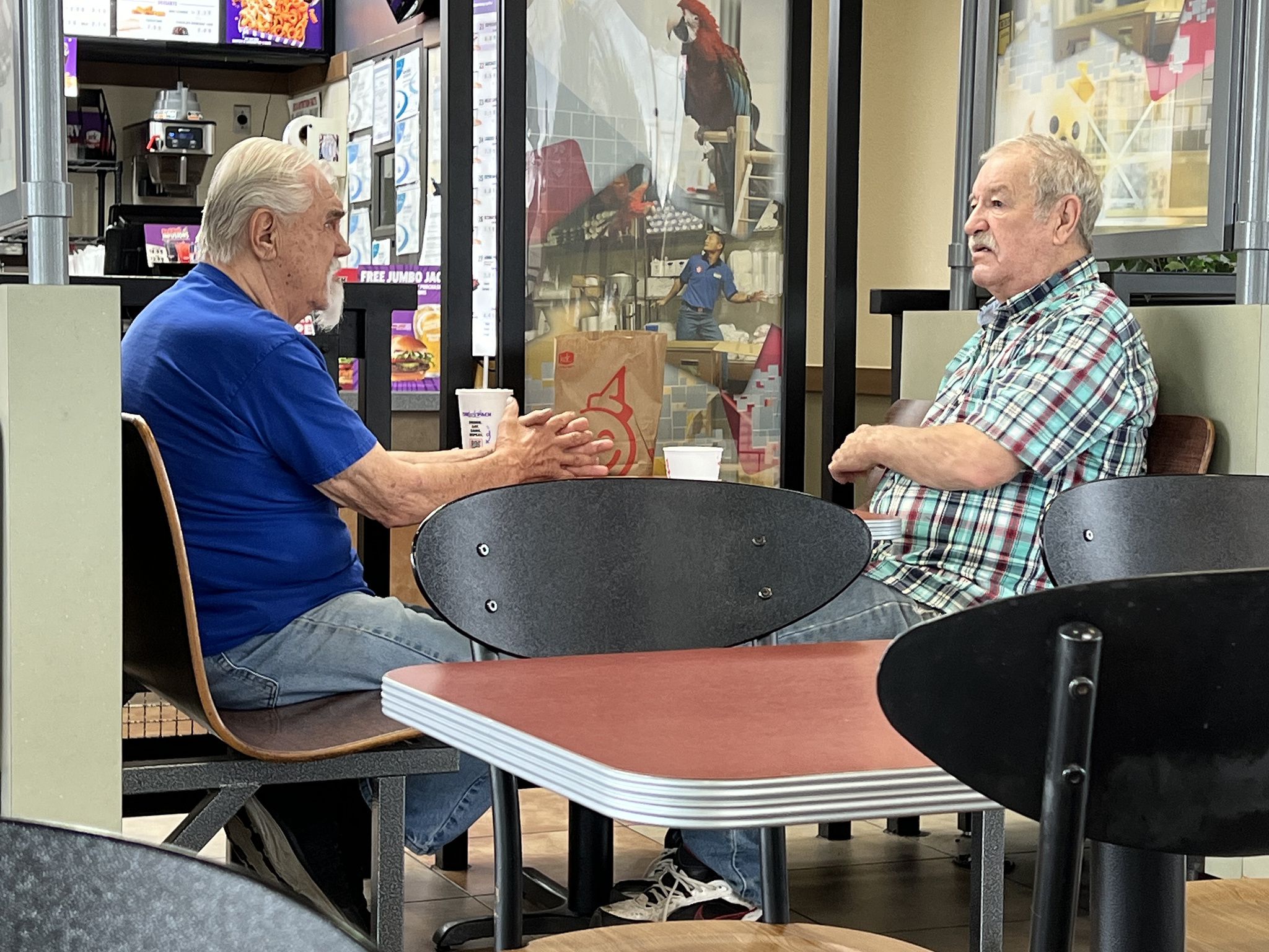 Two older men seated at a table in a fast food resturant. They have food in front of them, but are both staring off to the side of one another.