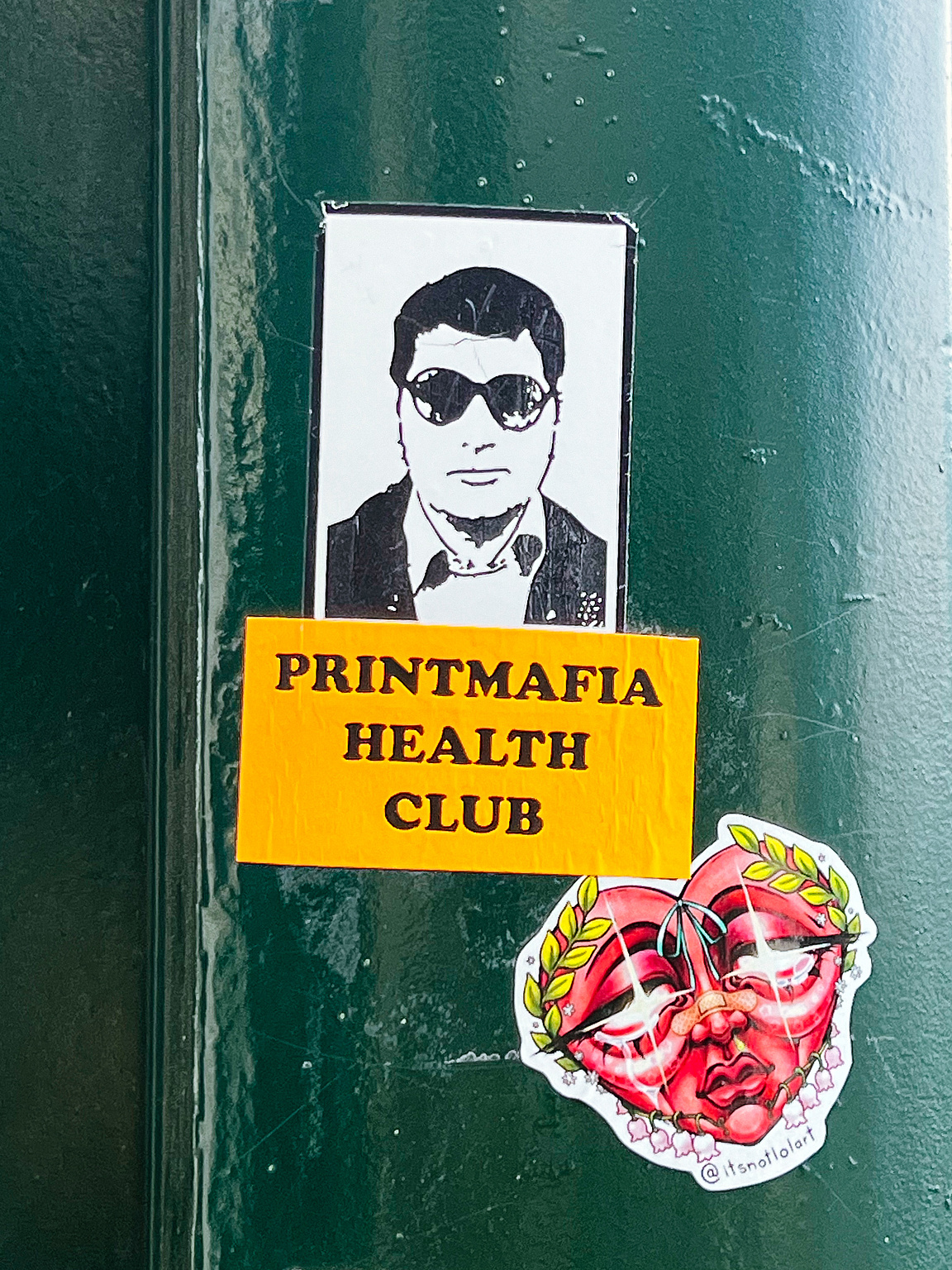 Stickers on a green surface: top, a black-and-white portrait of a man with sunglasses; middle, a yellow text reading "PRINTMAFIA HEALTH CLUB"; bottom, a colorful heart-shaped face sticker.